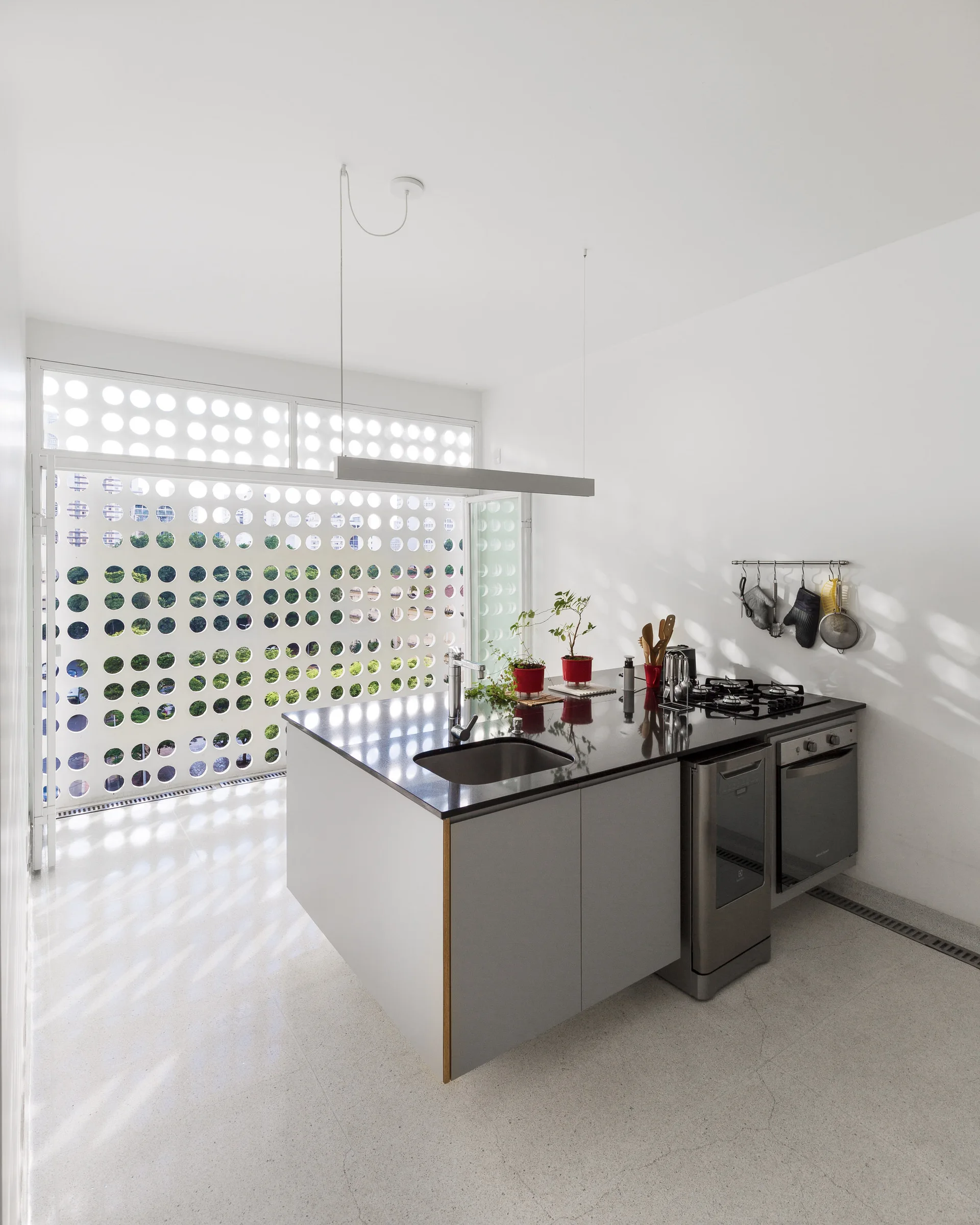 A minimalist white kitchen features a central grey island with a dark countertop and a large window covered by a perforated screen with circular cutouts.