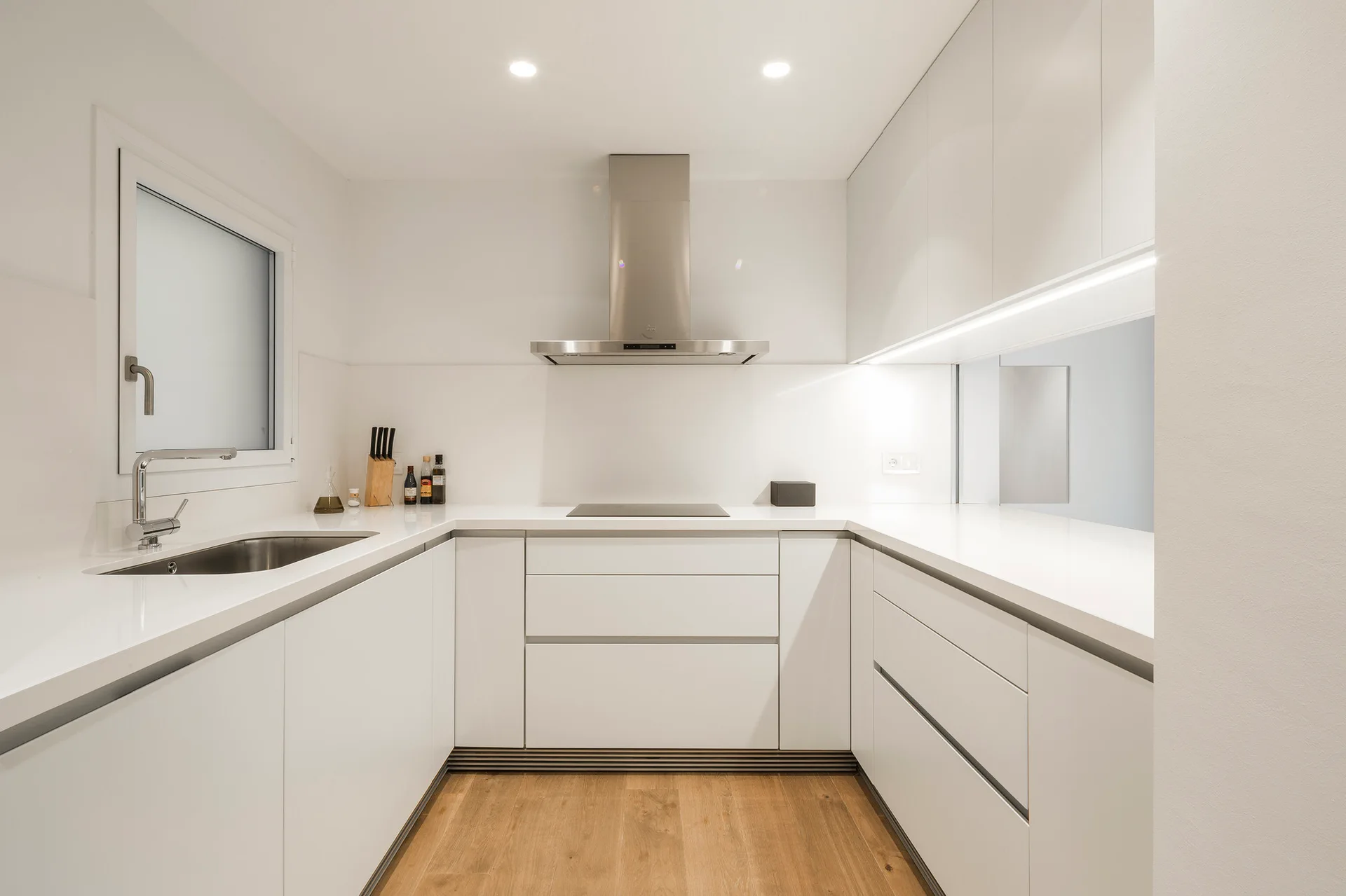 A minimalist U-shaped kitchen with flat-front white cabinets, a stainless steel range hood, and light hardwood flooring.