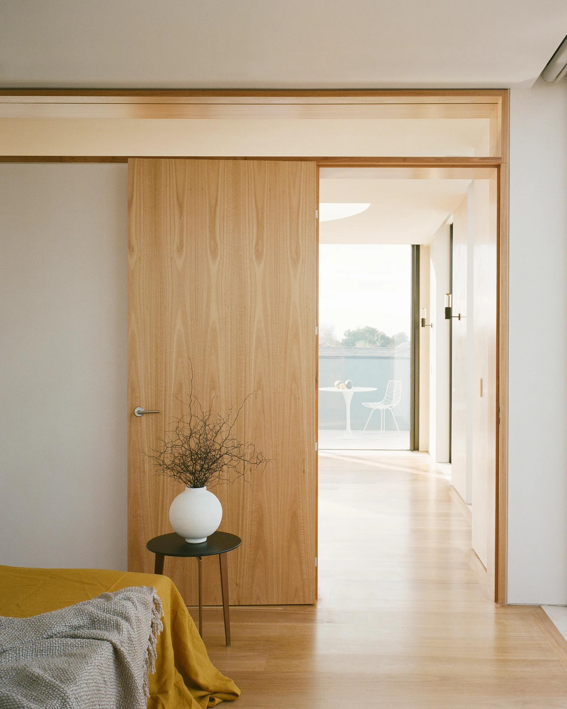 A minimalist bedroom features a large wood sliding door opening to a sunlit outdoor terrace with a white table and chair.