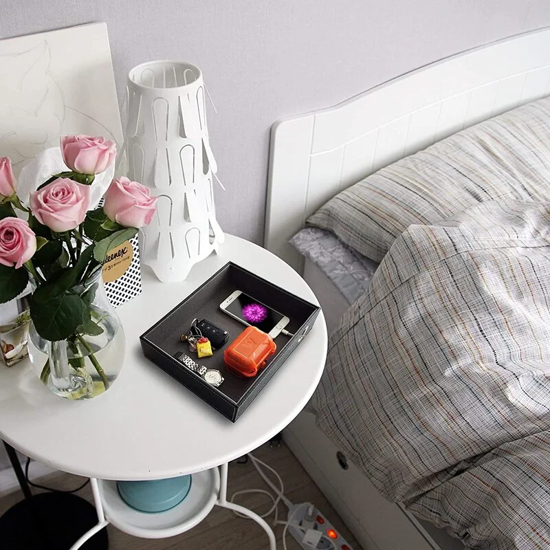 A round white nightstand beside a bed features a white sculptural lamp, a glass vase of pink roses, and a dark valet tray with personal items.