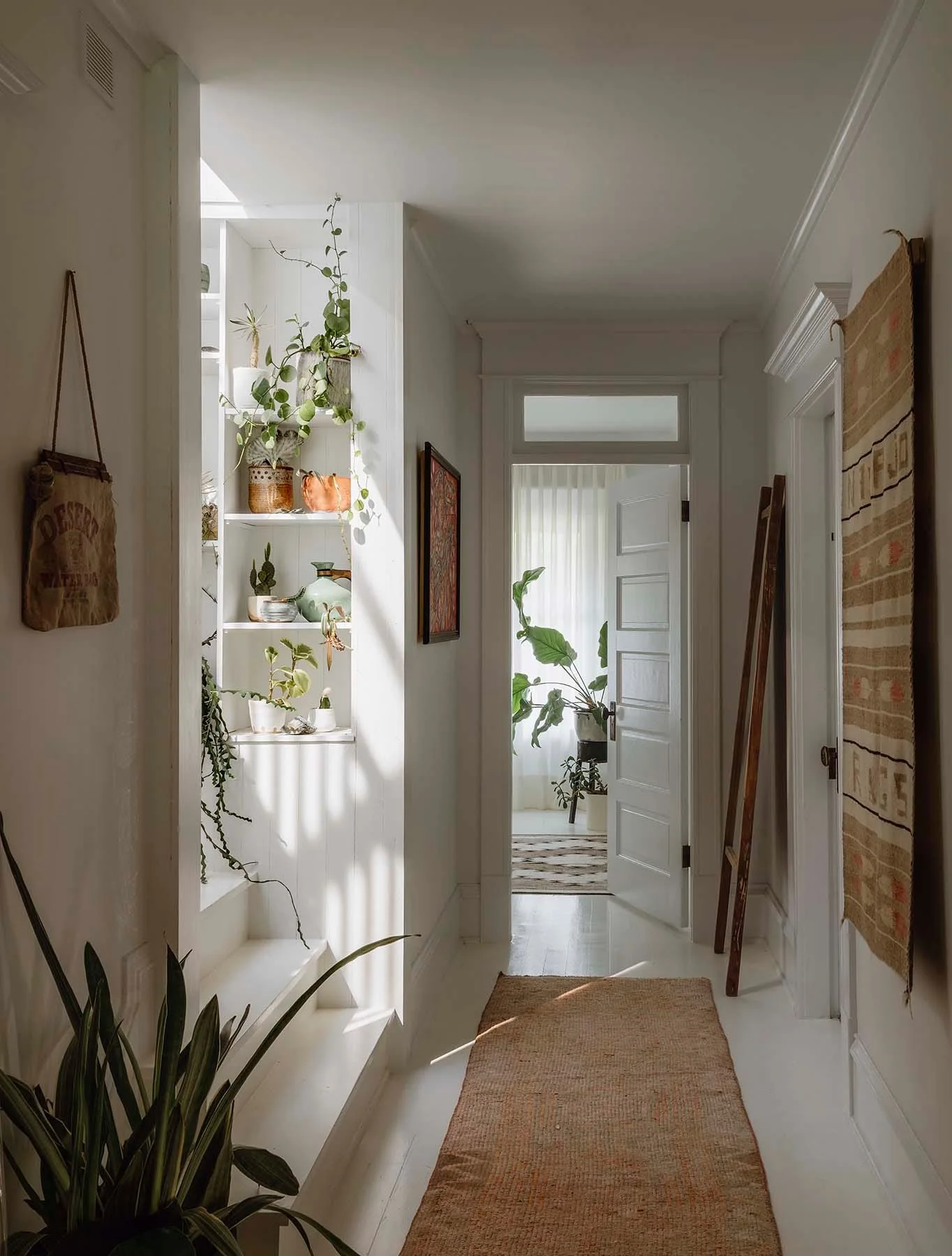 A bright white hallway features built-in plant shelves, a wooden ladder, a woven wall hanging, and a natural fiber runner rug.