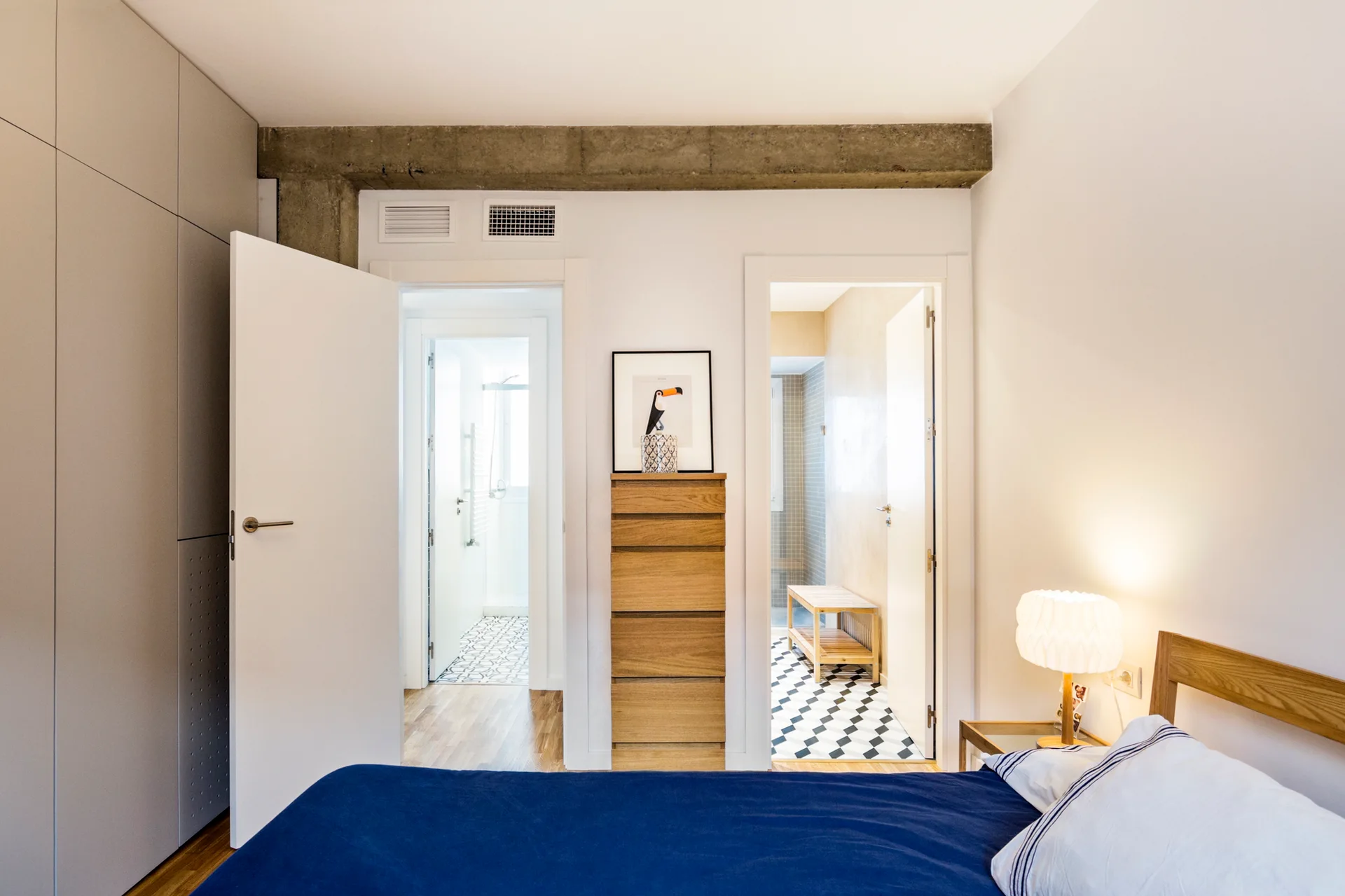 A minimalist bedroom featuring a blue bed, a tall wooden dresser, grey built-in closets, and an exposed concrete beam above two open bathroom doors.