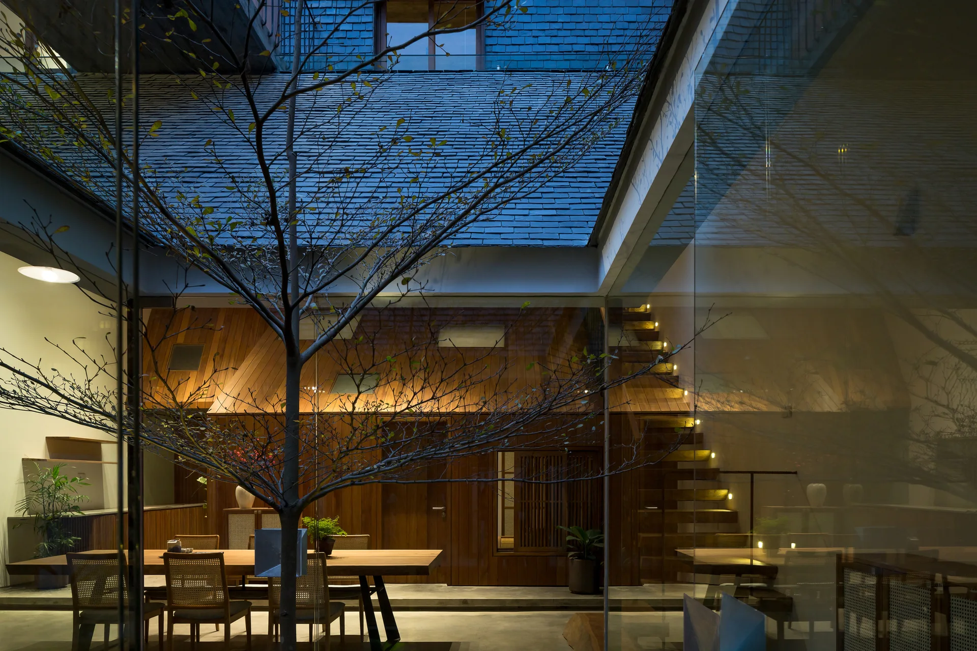 A modern dining room with a long wood table and cane chairs is viewed through a glass partition next to an indoor tree and a wooden staircase.