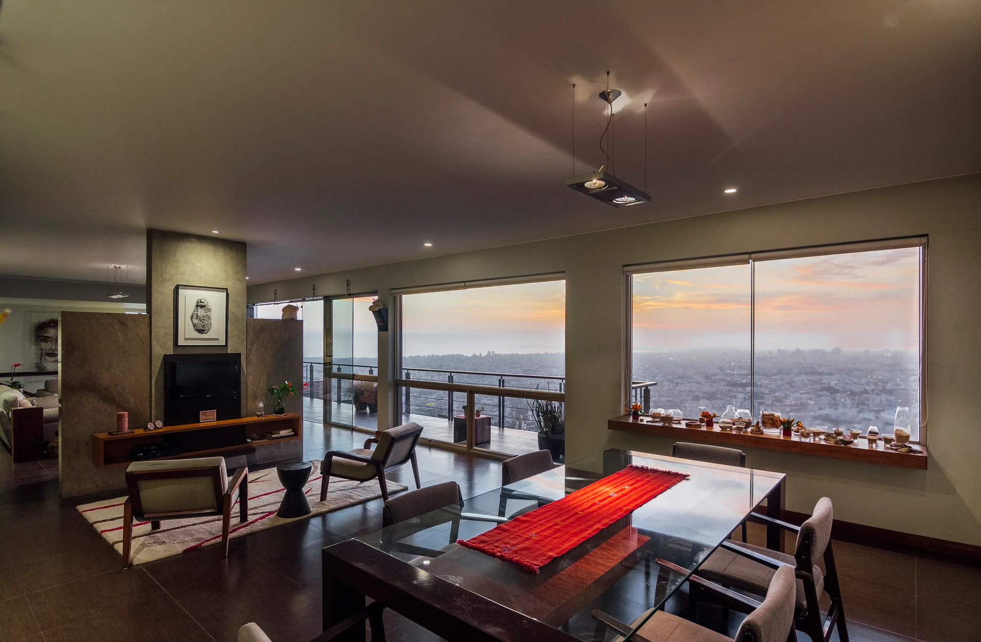 An open-concept living and dining area with a glass-topped table, a stone-textured partition wall, and large windows showcasing a city sunset.