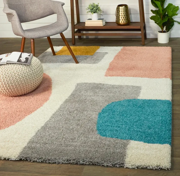 A multicolored shag area rug with bold geometric blocks of teal, pink, and gray sits on a dark wood floor near a gray armchair and knit pouf.
