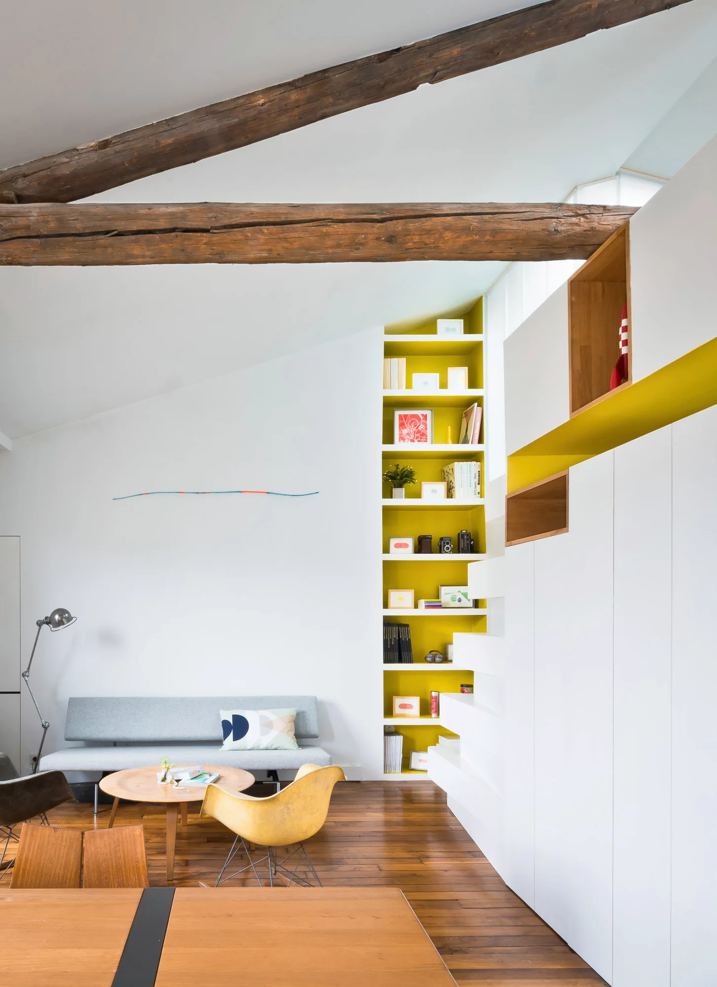 A living room with exposed wood ceiling beams features a grey sofa, a round wooden coffee table, and a tall recessed shelving unit with a yellow interior.