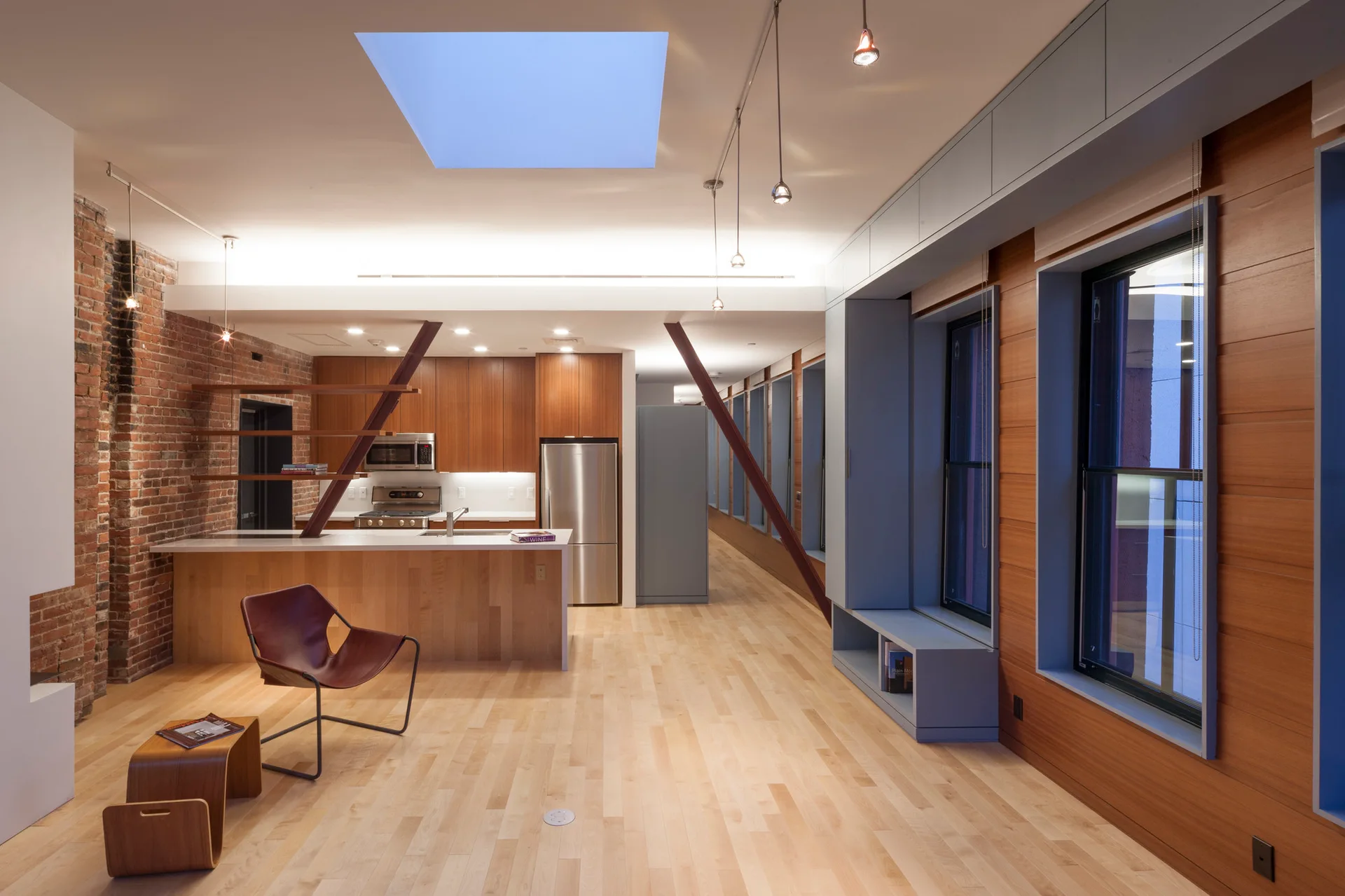 An open loft kitchen and living area featuring a central wood island, diagonal structural steel beams, exposed brick, and a large ceiling skylight.