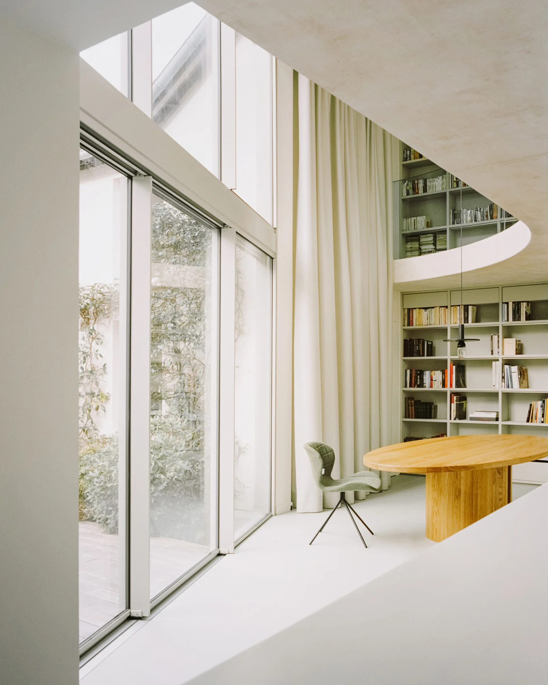 A double-height home library features an oval wooden table, a light green chair, floor-to-ceiling windows, and a curved mezzanine with built-in bookshelves.