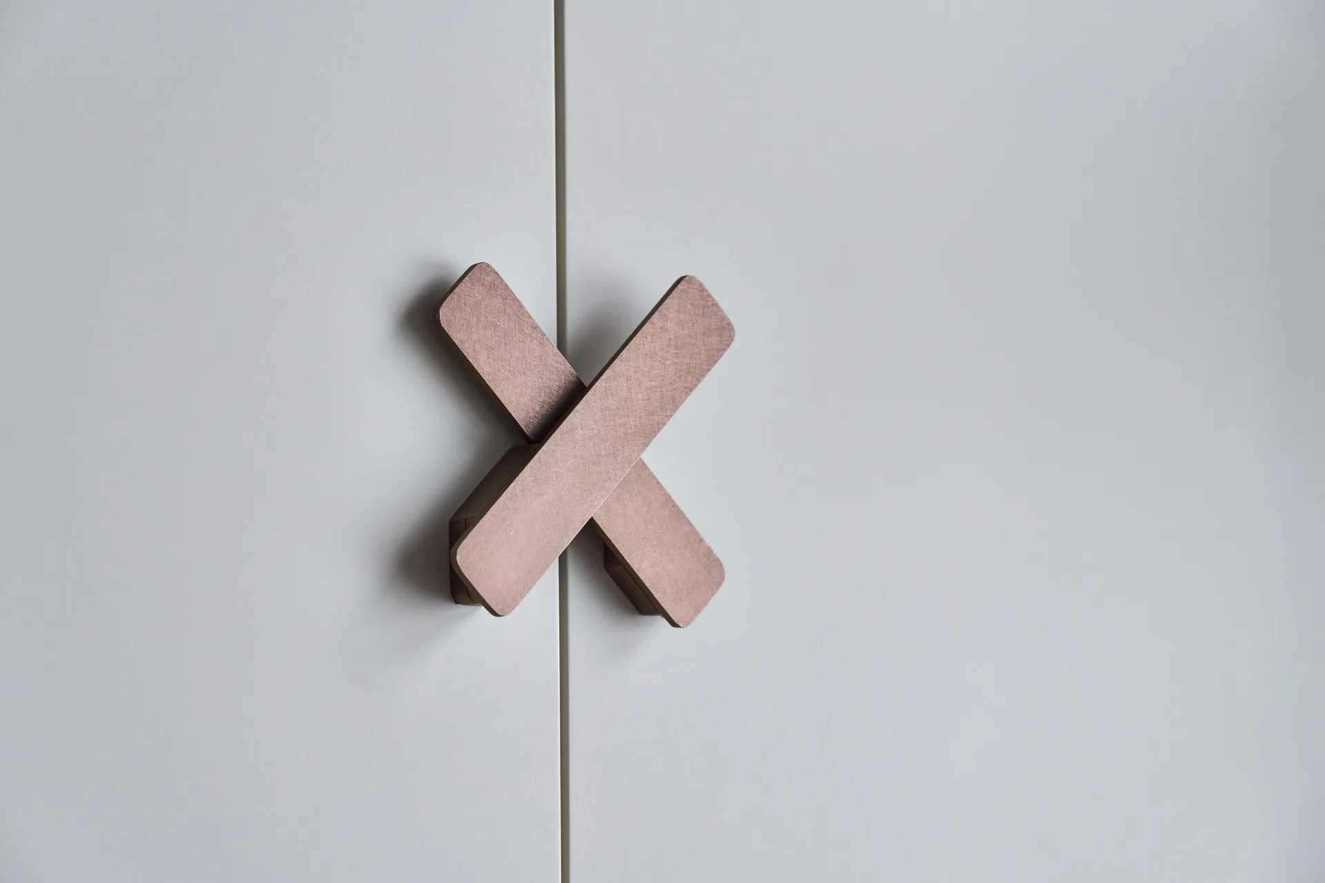 A close-up of a brushed copper X-shaped metal handle mounted on flat-panel white matte wardrobe doors.