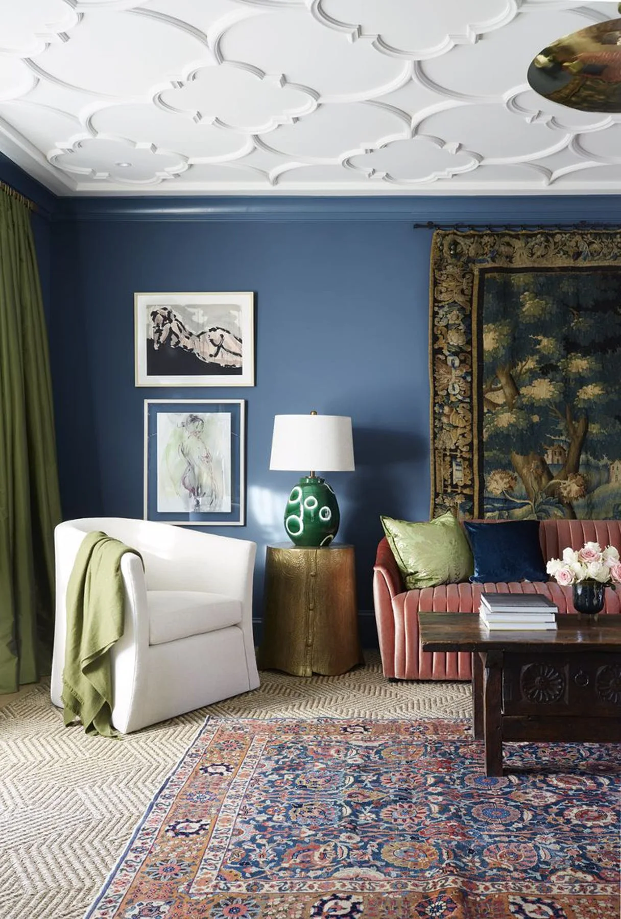 An eclectic living room with deep blue walls, a white ornamental relief ceiling, a pink channel-tufted sofa, and a green ceramic table lamp.