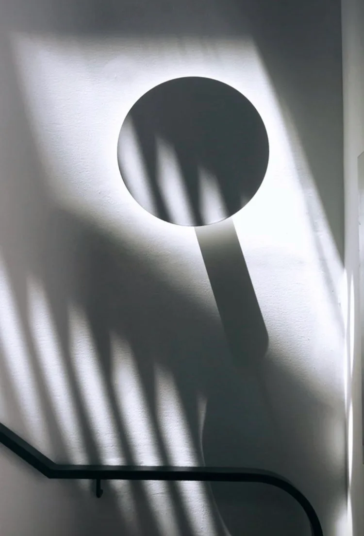 A backlit circular wall sconce casts a soft halo on a white staircase wall decorated with geometric shadows from a black handrail.