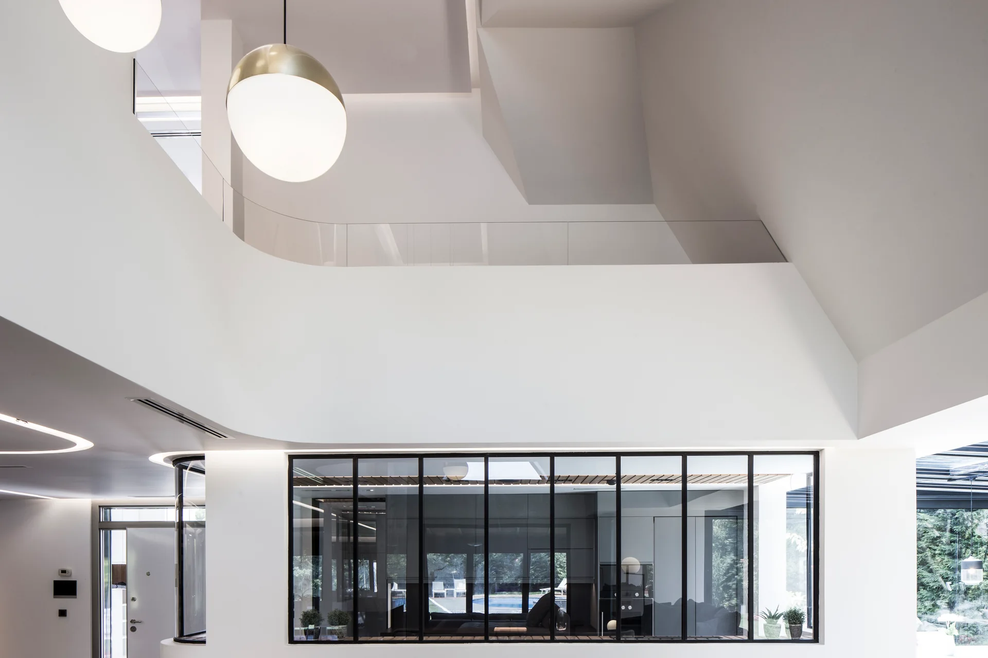 A minimalist double-height residential interior featuring white walls, a frameless glass balcony railing, and a large black-framed glass partition wall.