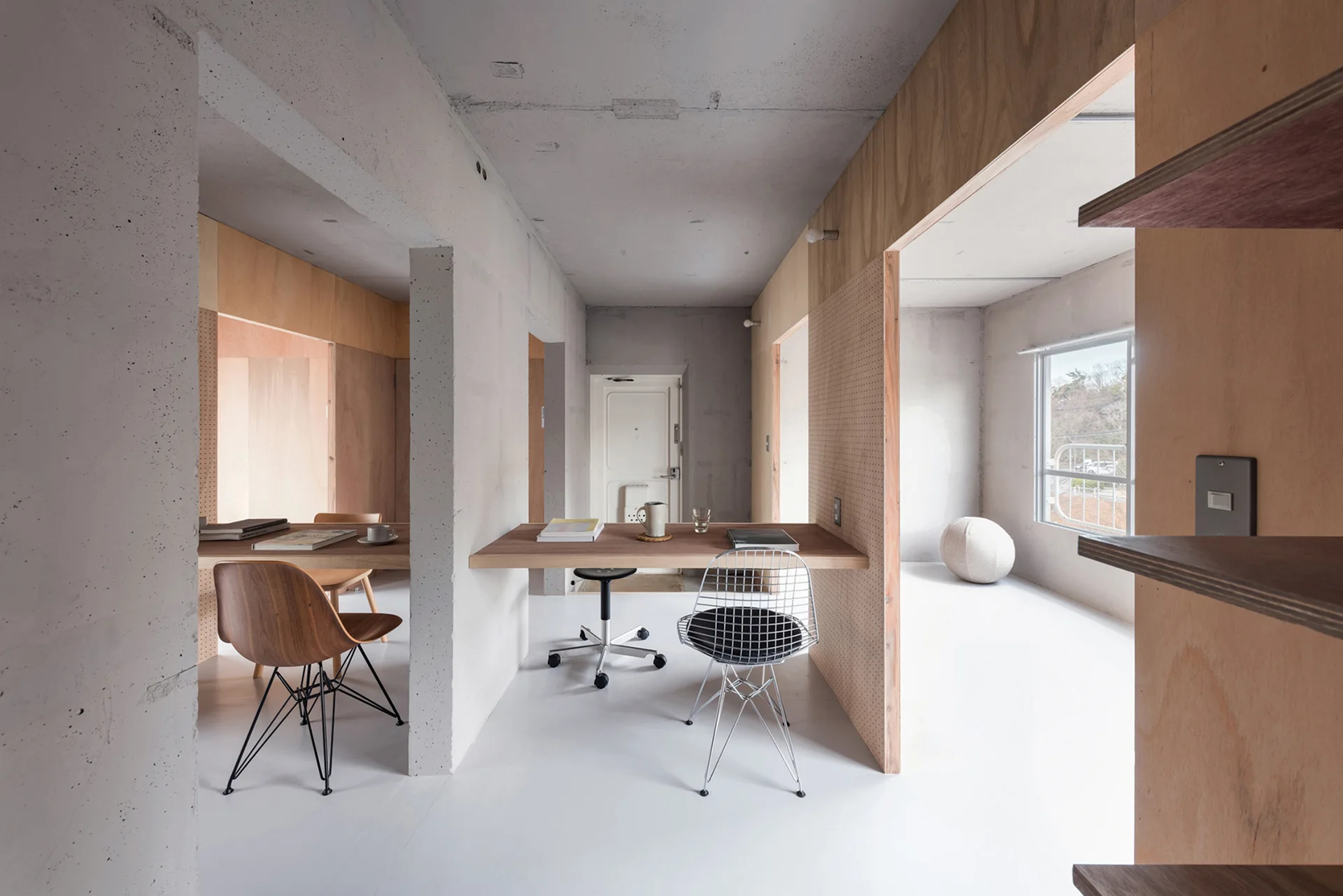 A minimalist home office featuring raw concrete walls, warm plywood partitions, and wooden floating desks on a white floor.