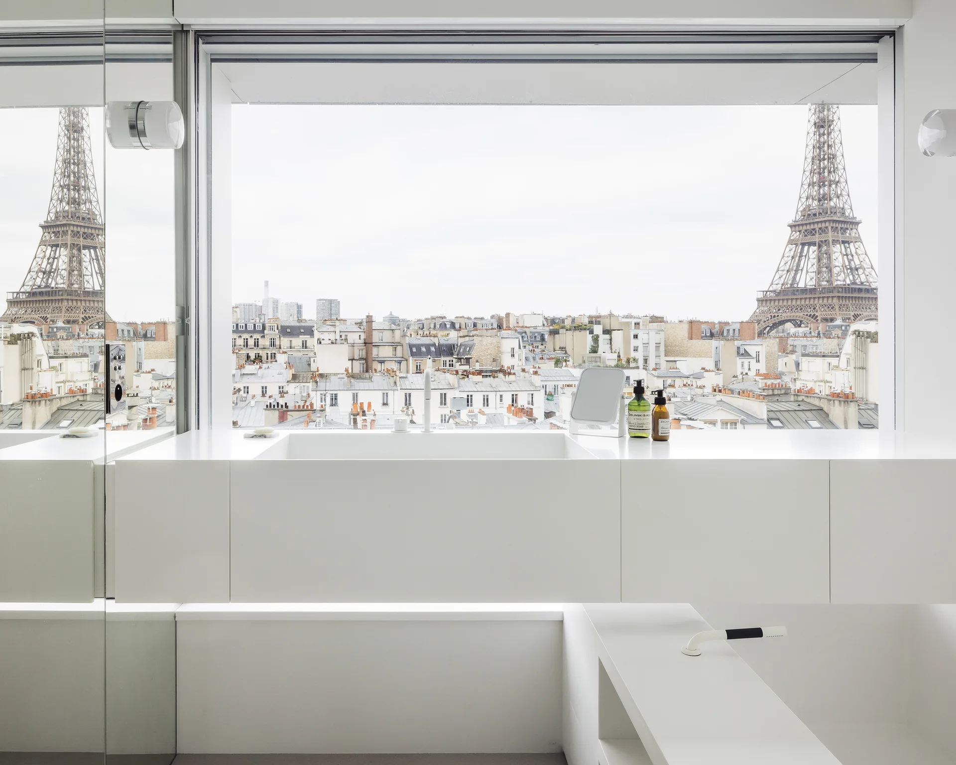 A minimalist white vanity with an integrated sink is set against a large picture window showing a view of the Eiffel Tower in Paris.