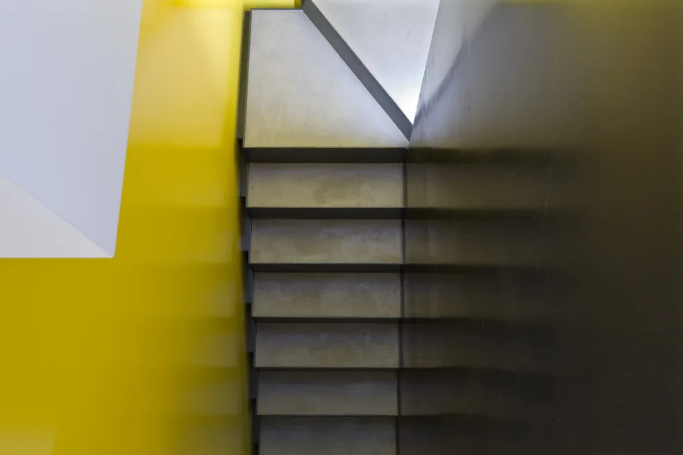 An overhead view of minimalist concrete stairs bordered by a bright yellow wall and a dark glossy wall.