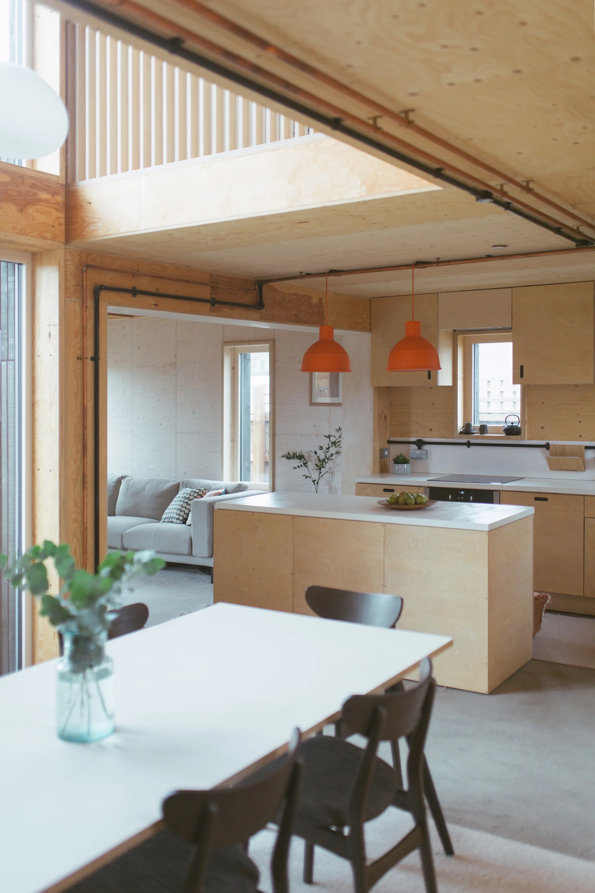 A modern open-plan kitchen and dining area featuring light plywood walls and cabinetry, a central island, and two bright orange pendant lights.
