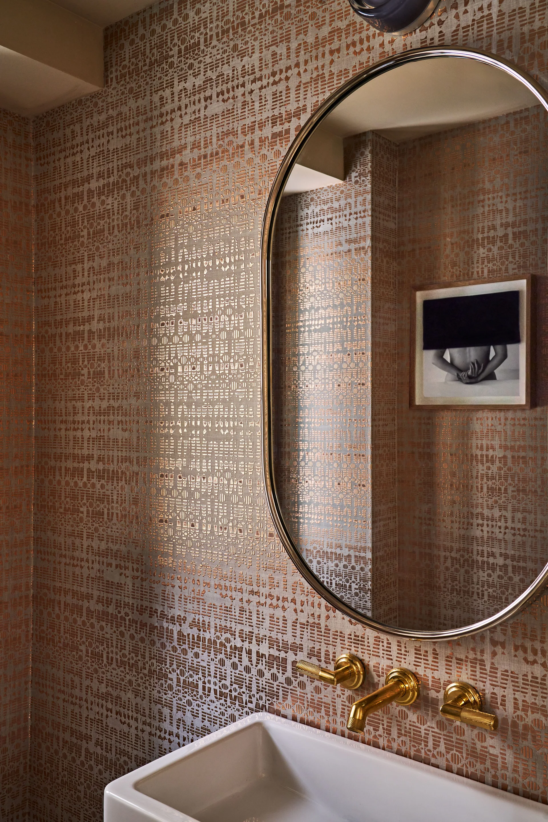 A compact powder room with metallic copper geometric wallpaper, a gold wall-mounted faucet over a white sink, and an oval mirror.