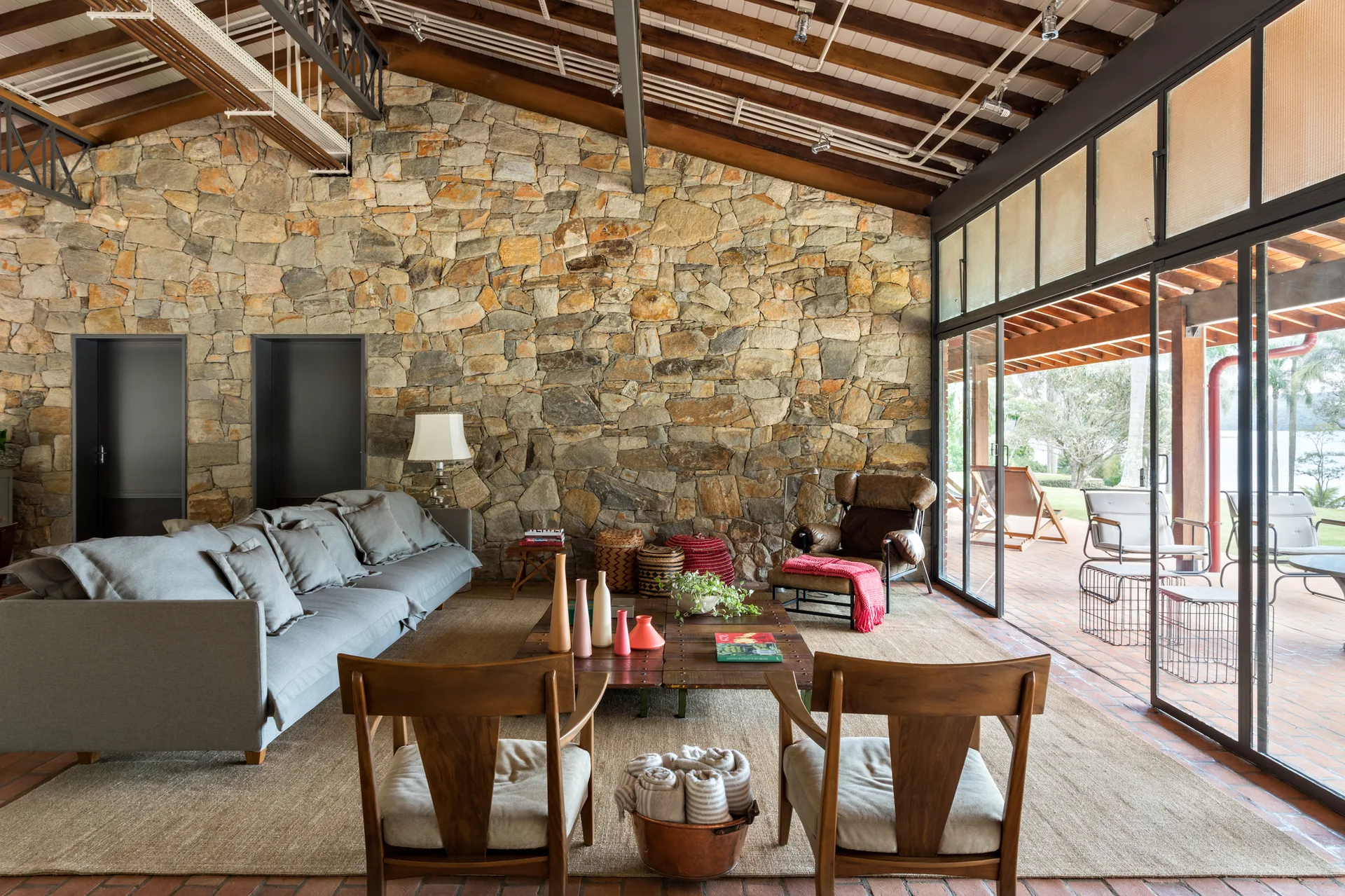 A living room featuring a rough stone accent wall, exposed wooden ceiling beams, a grey sofa, and large glass sliding doors leading to a patio.