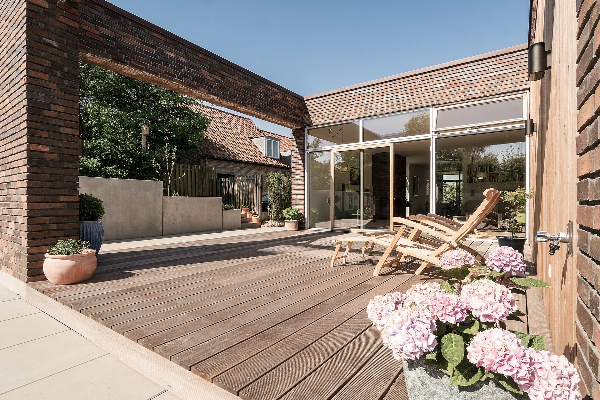 A sunny wooden deck patio features reclining lounge chairs next to large glass doors and red brick walls with pink hydrangeas in the foreground.