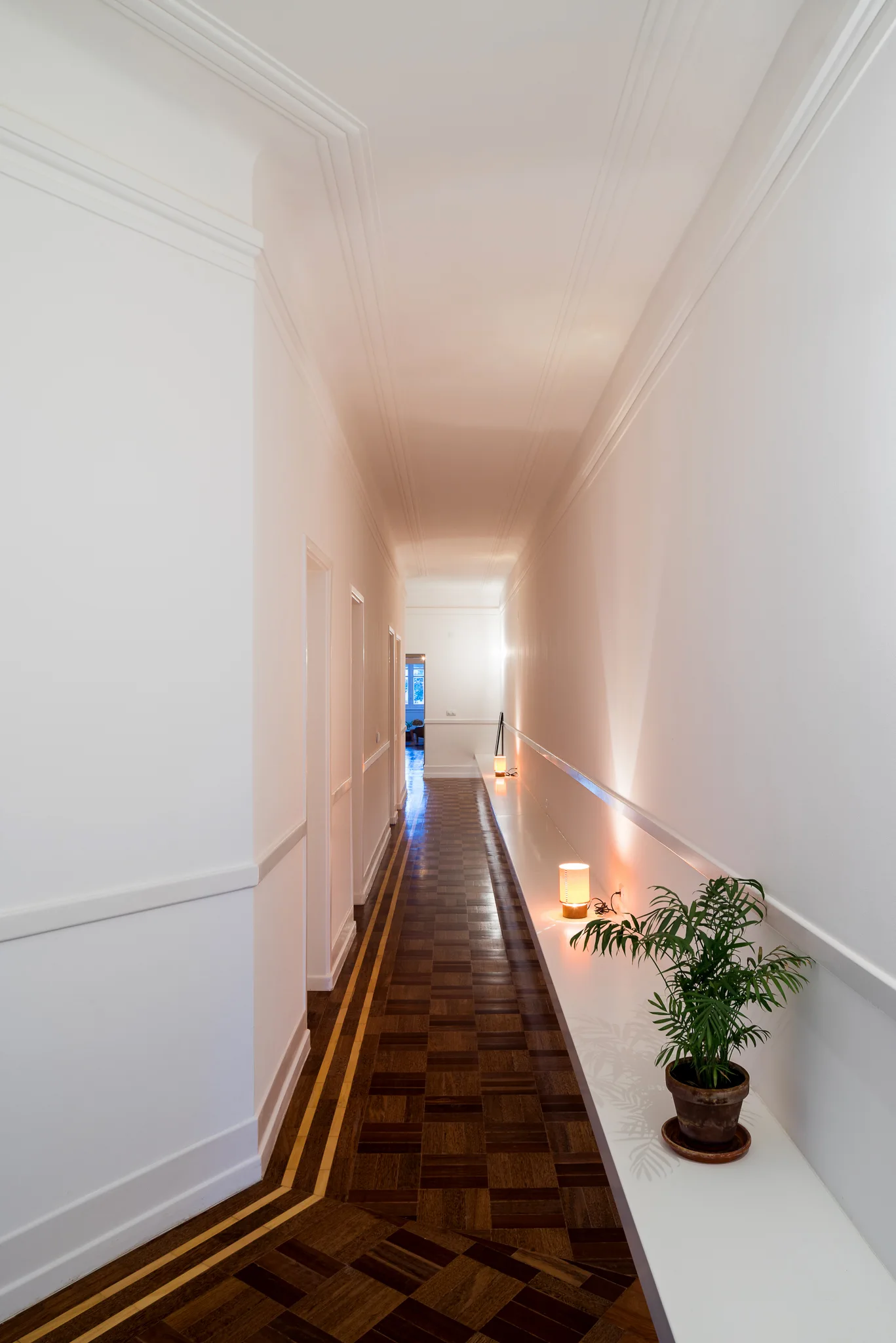 A long white hallway features geometric parquet wood flooring, elegant wall wainscoting, and a low built-in console shelf.