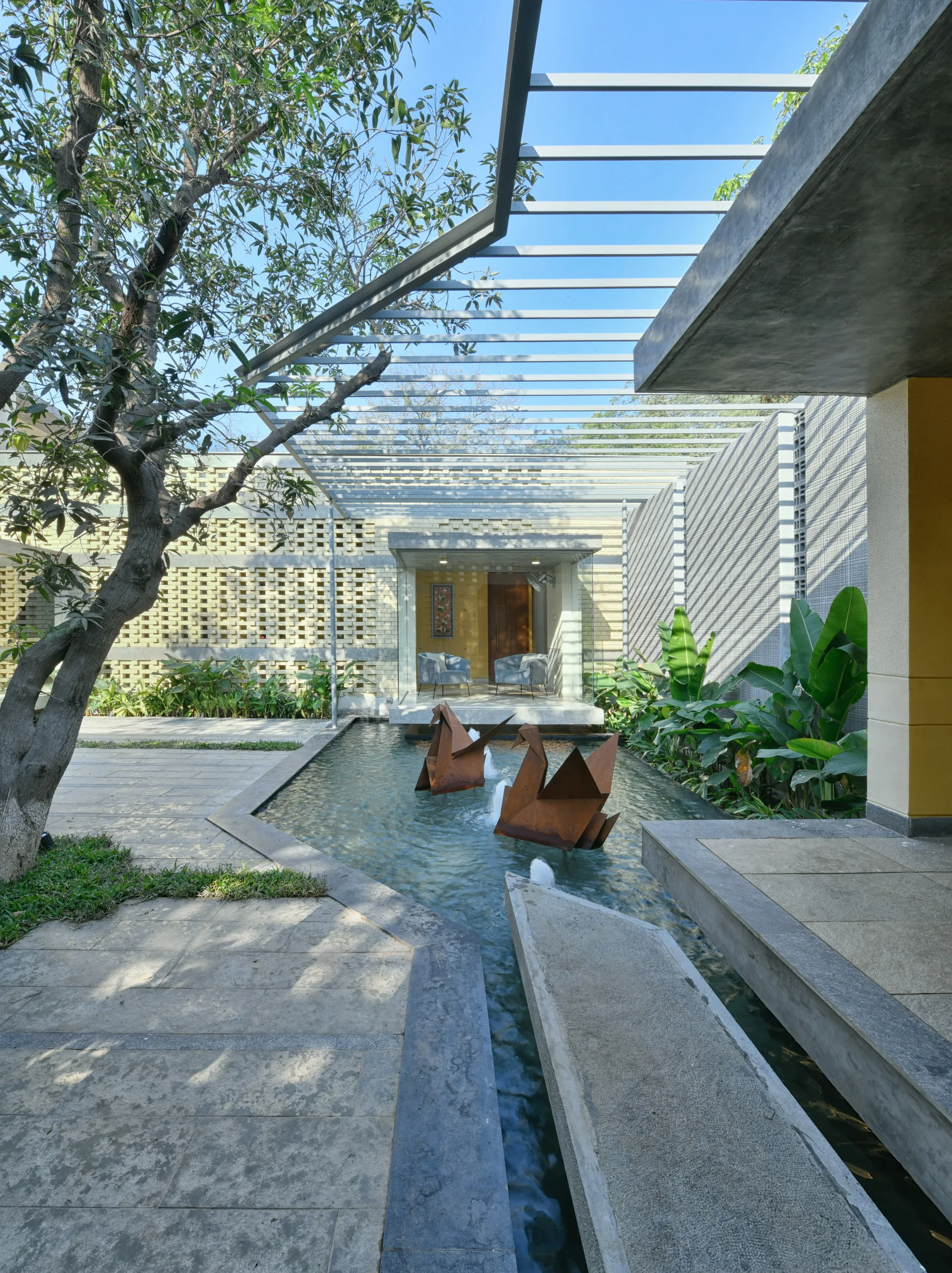 A modern outdoor courtyard featuring a stone-paved reflecting pool with rusted metal bird sculptures under a geometric steel pergola.