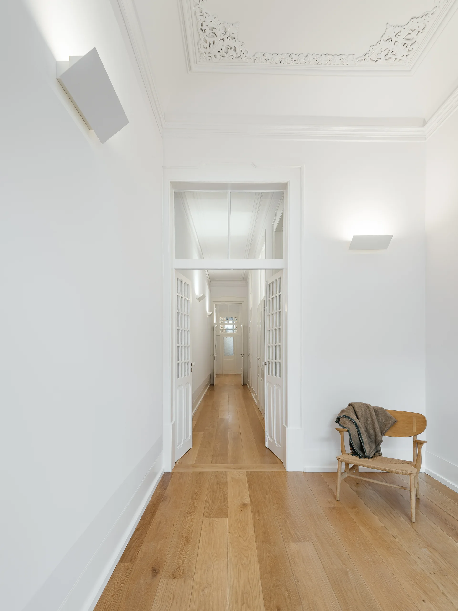 A bright white hallway with light hardwood floors, detailed plaster ceiling molding, open glass-paned doors, and a wooden chair.