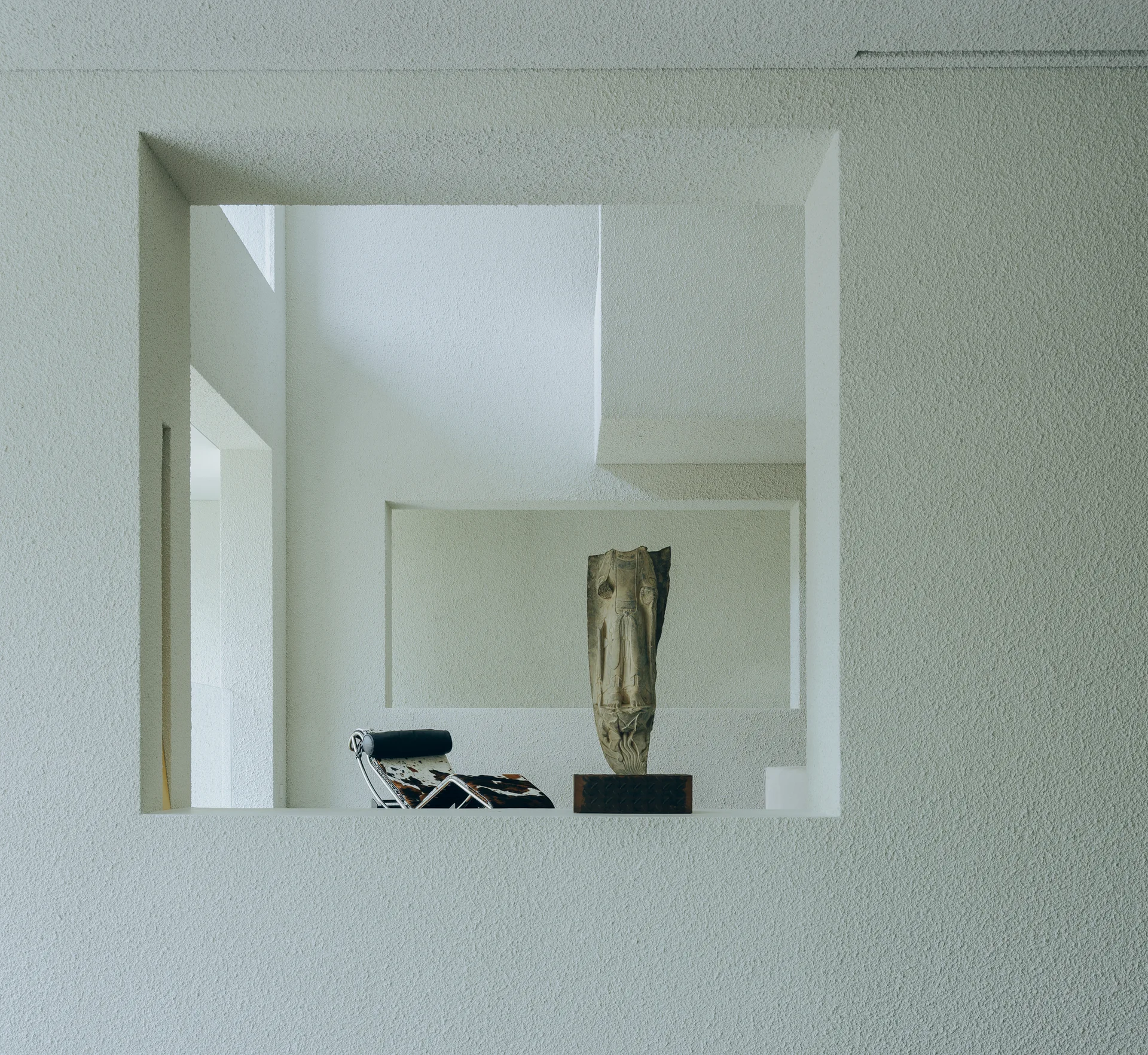 An architectural cutout in a rough off-white stucco wall reveals a modernist cowhide chaise lounge and a stone sculpture on a dark wood pedestal.