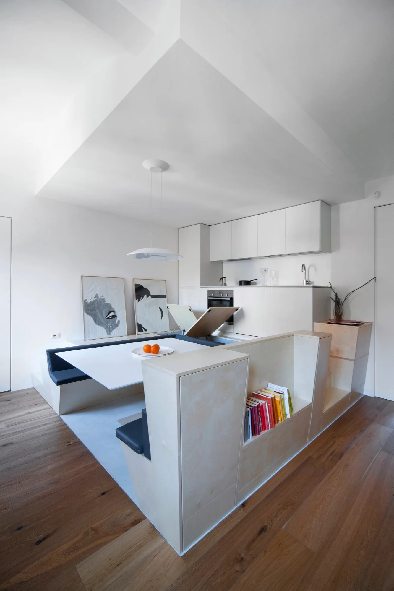 A compact kitchen with flat-front white cabinets features a custom plywood dining nook with built-in benches, a white table, and an integrated outer bookshelf.
