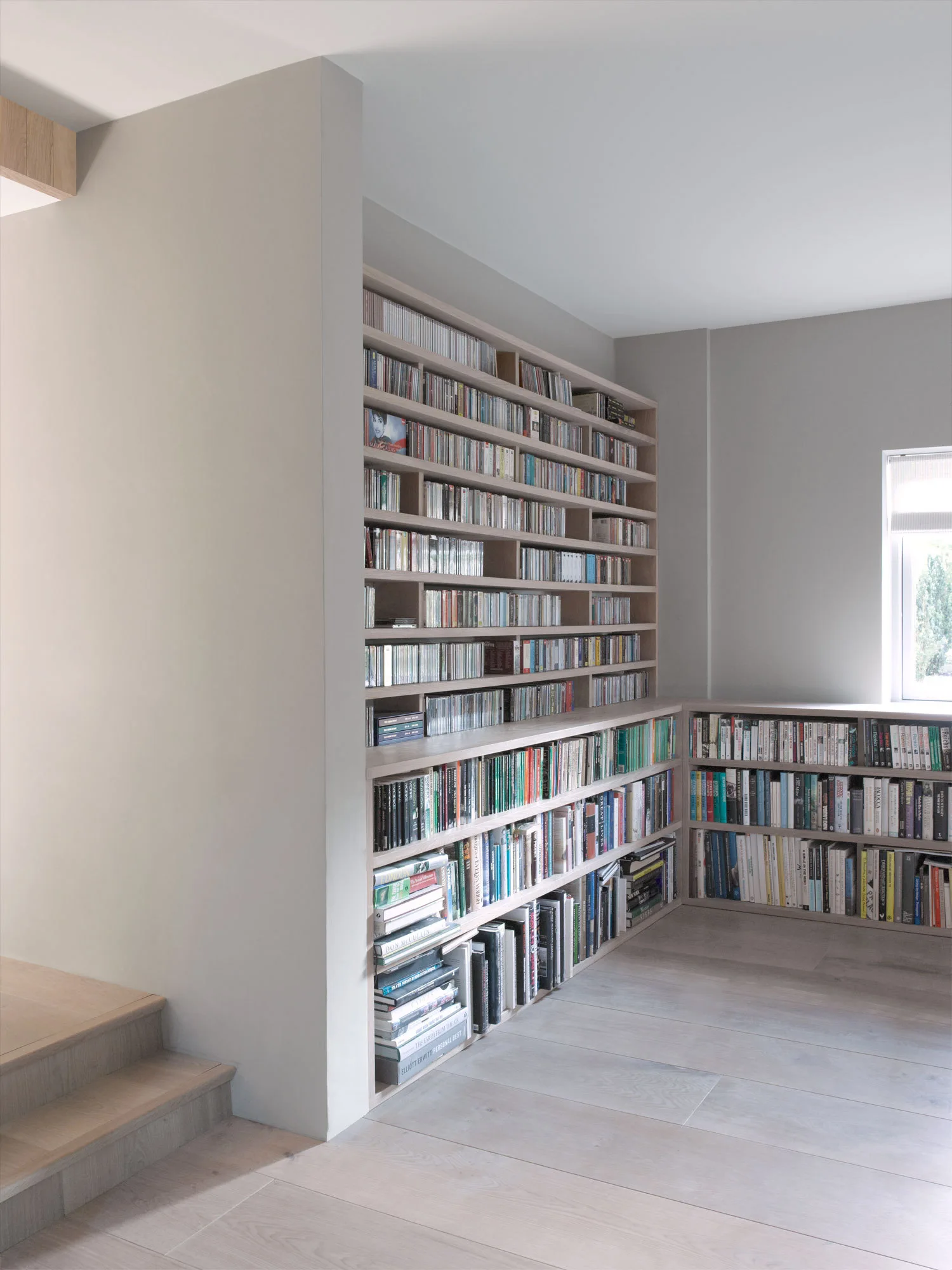A minimalist library space with custom light wood corner bookshelves filled with books and CDs, featuring neutral grey walls and matching light wood floors.