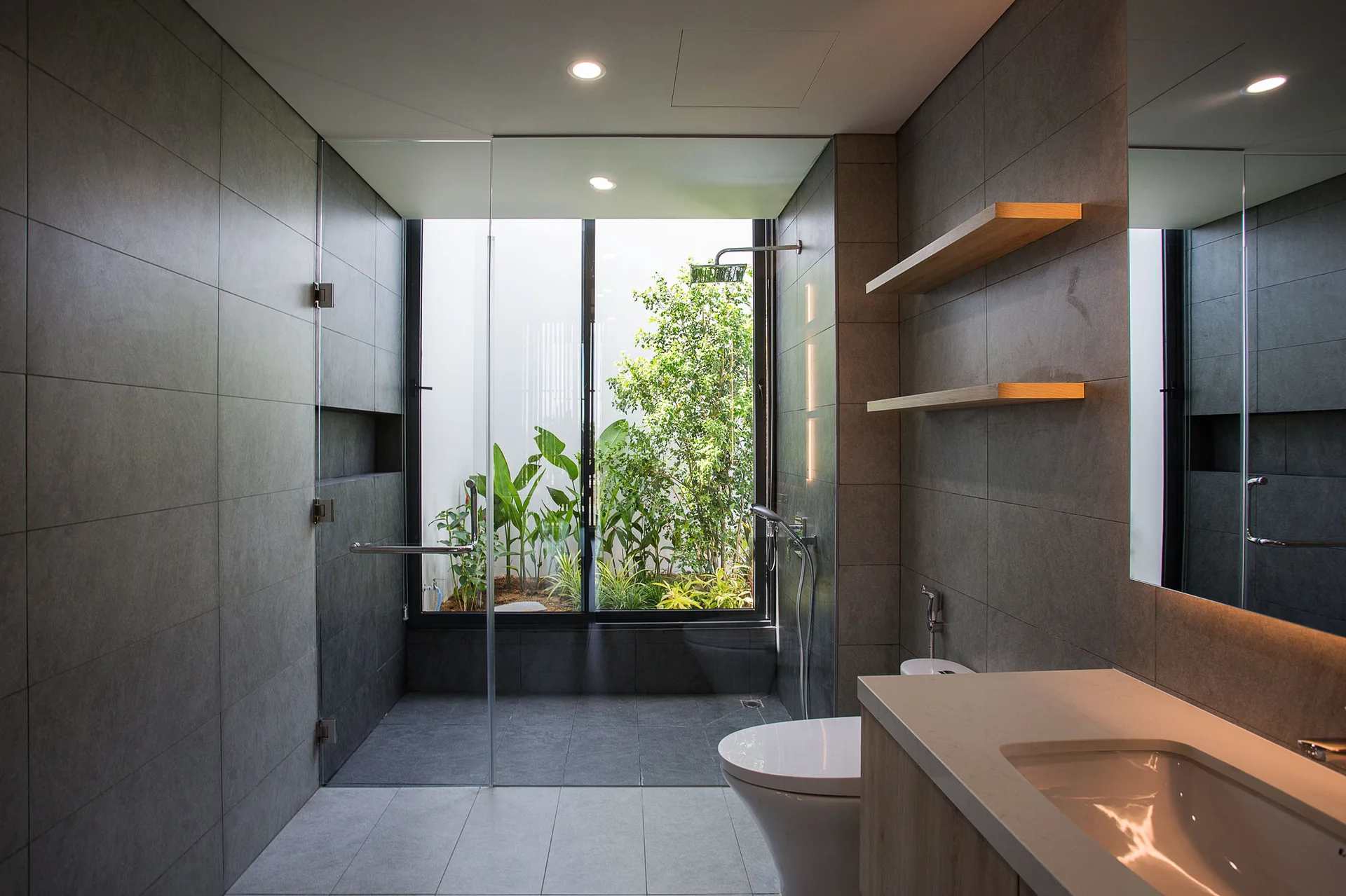A modern grey tiled bathroom features a glass walk-in shower looking out onto a garden, a toilet, floating shelves, and a wood vanity.