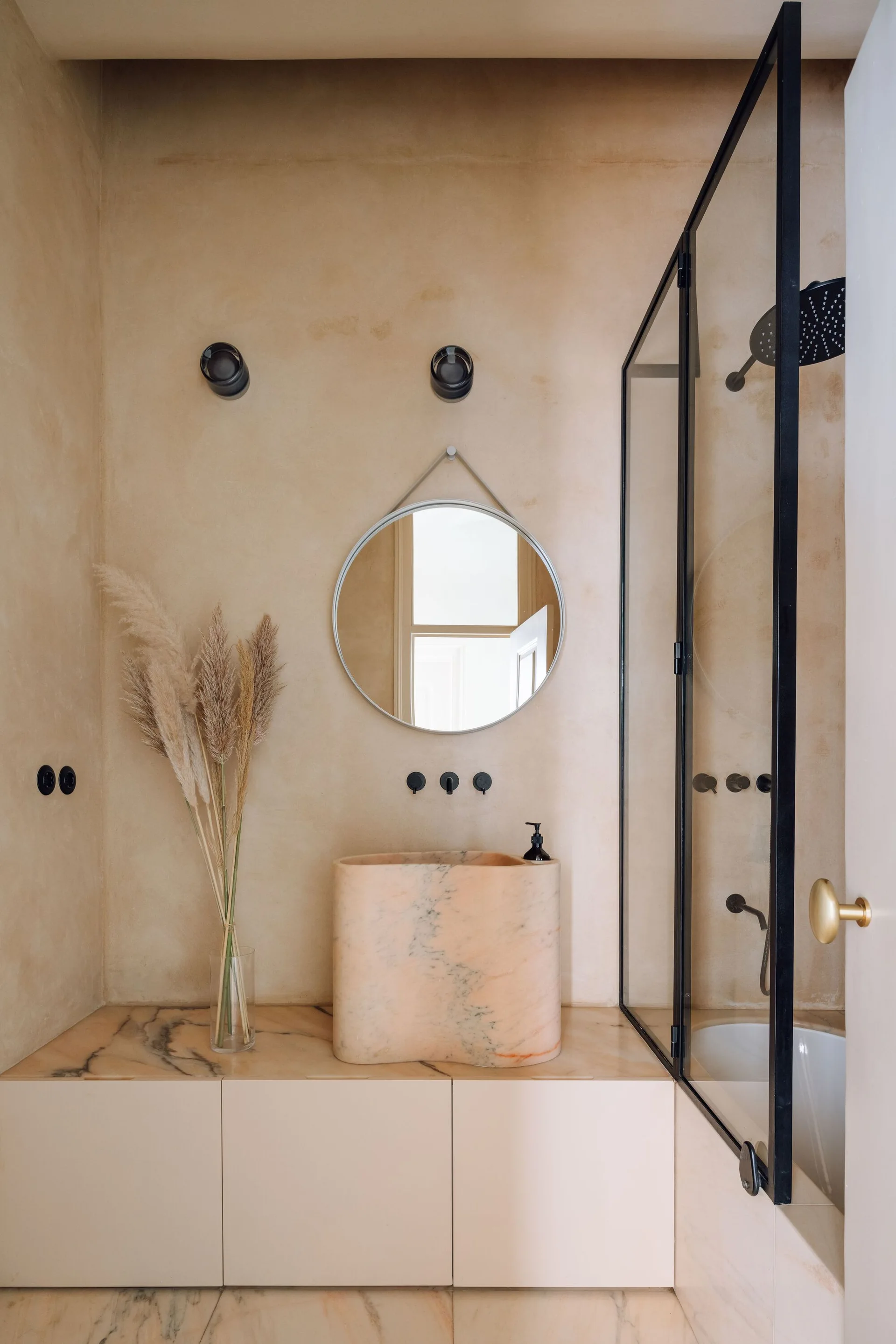 A minimalist bathroom features a sculptural peach-toned stone sink on a marble countertop beneath a round mirror and black wall-mounted faucet.