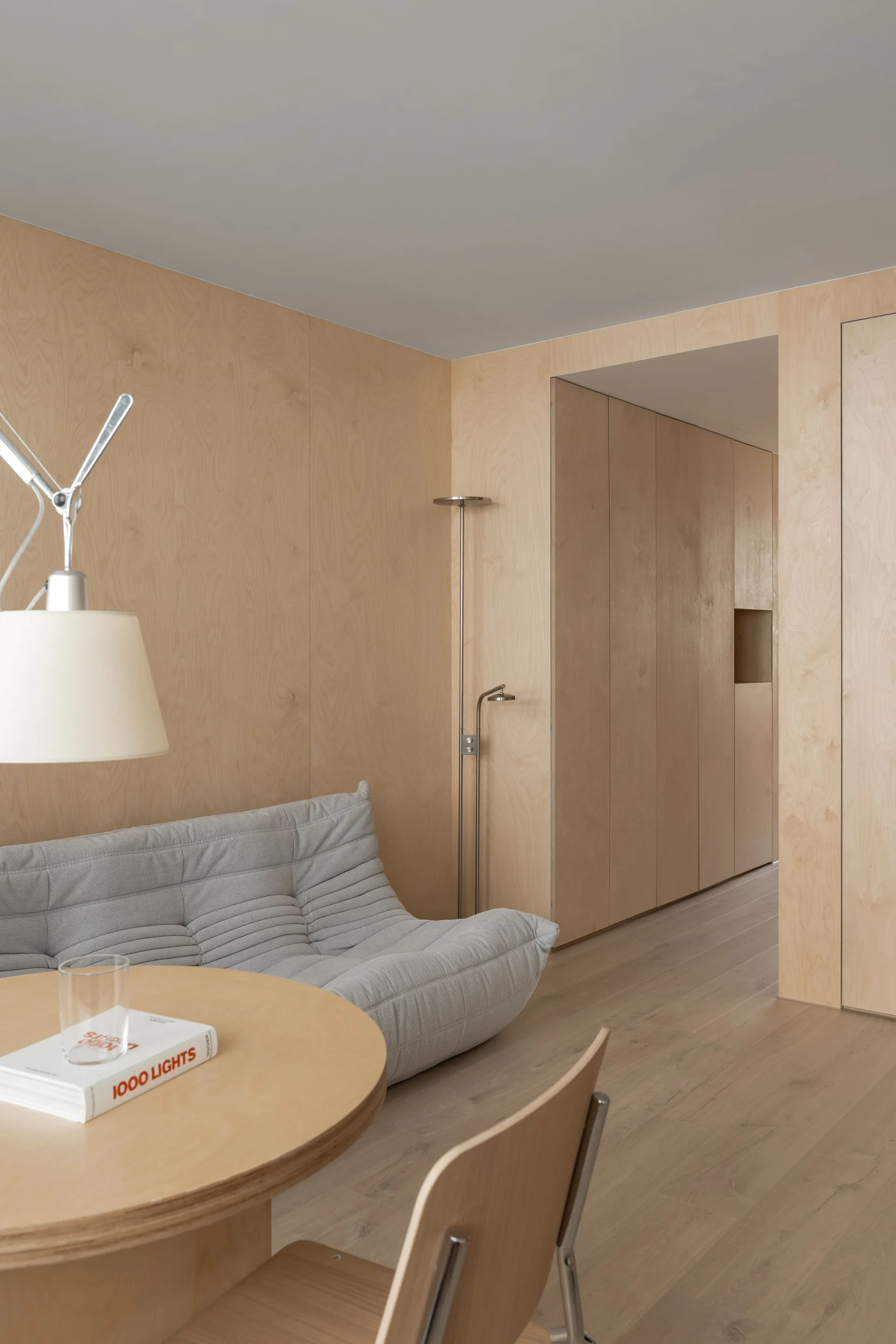 A minimalist living room features light birch plywood wall paneling, a low gray pleated sofa, a round wooden table, and integrated storage doors.