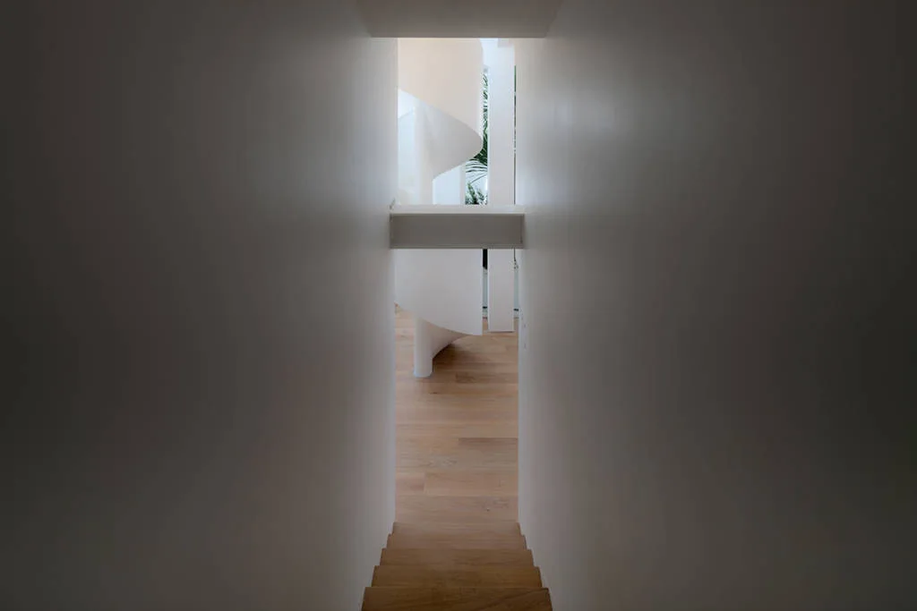 Looking through a narrow white corridor toward a sculptural white spiral staircase on light wood flooring.