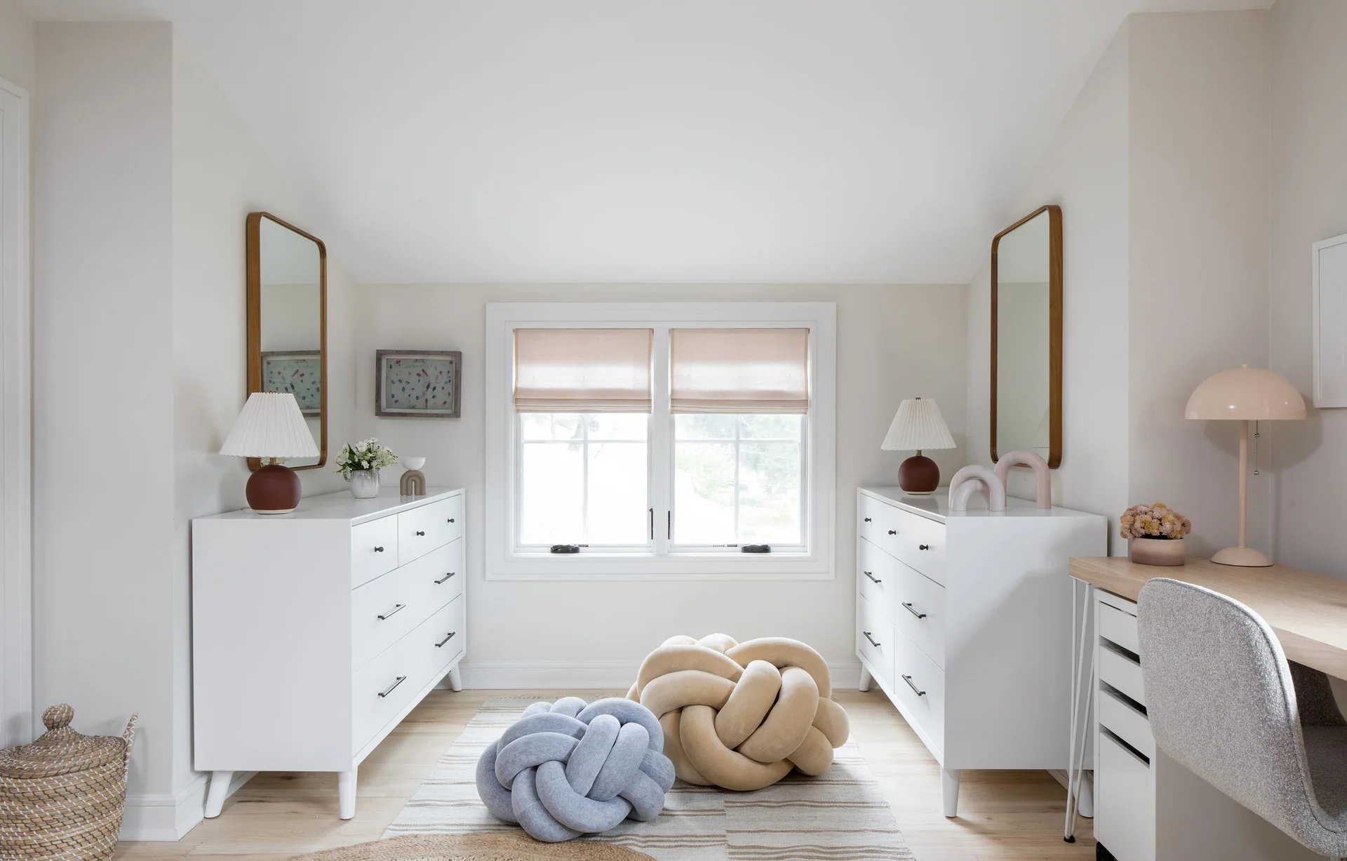 Two symmetrical white dressers with matching mirrors flank a window, with two large fabric knot pillows resting on a striped rug below.