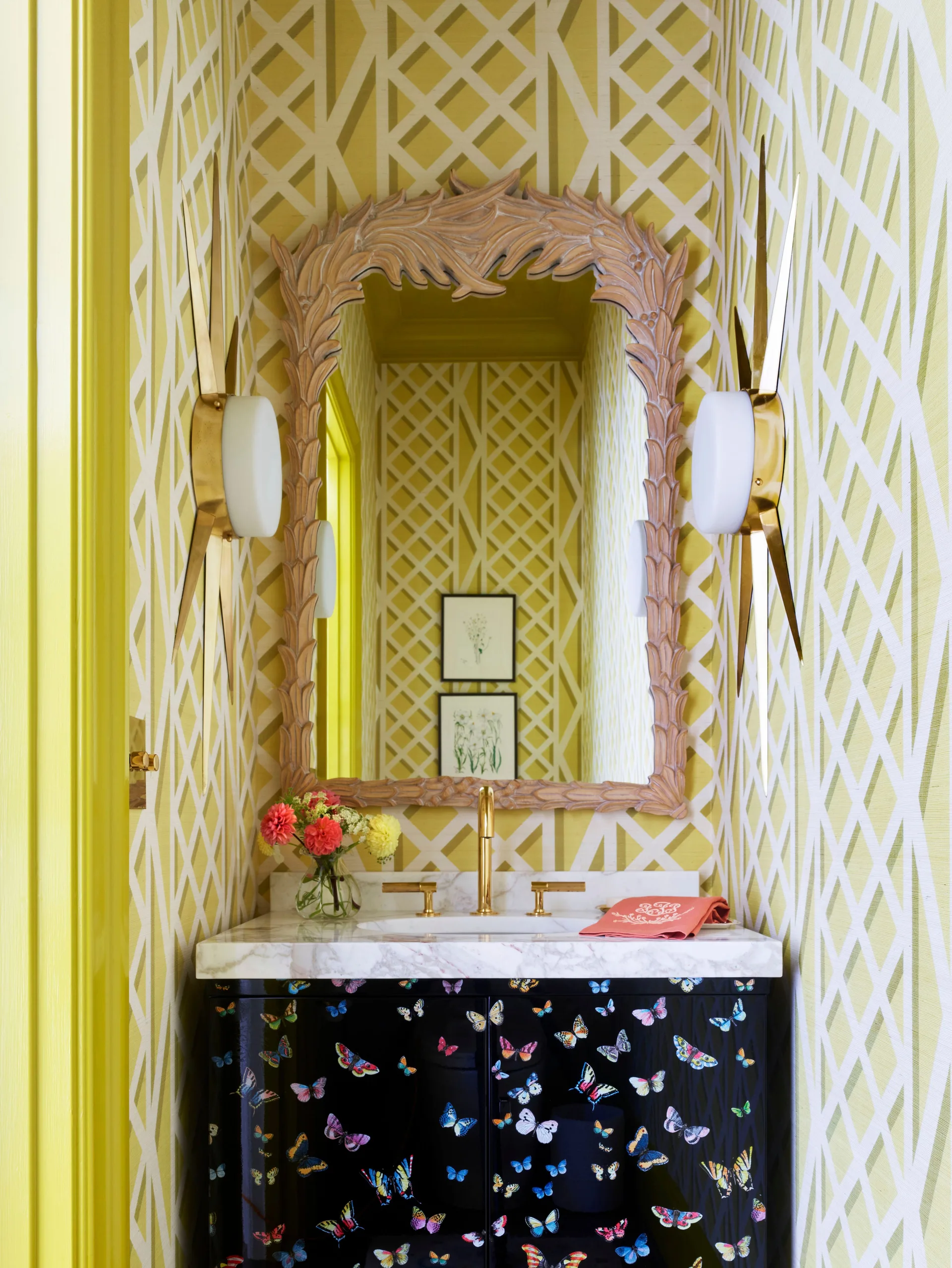 A bright yellow powder room with lattice wallpaper, an ornate wood mirror, starburst sconces, and a black butterfly-printed vanity.