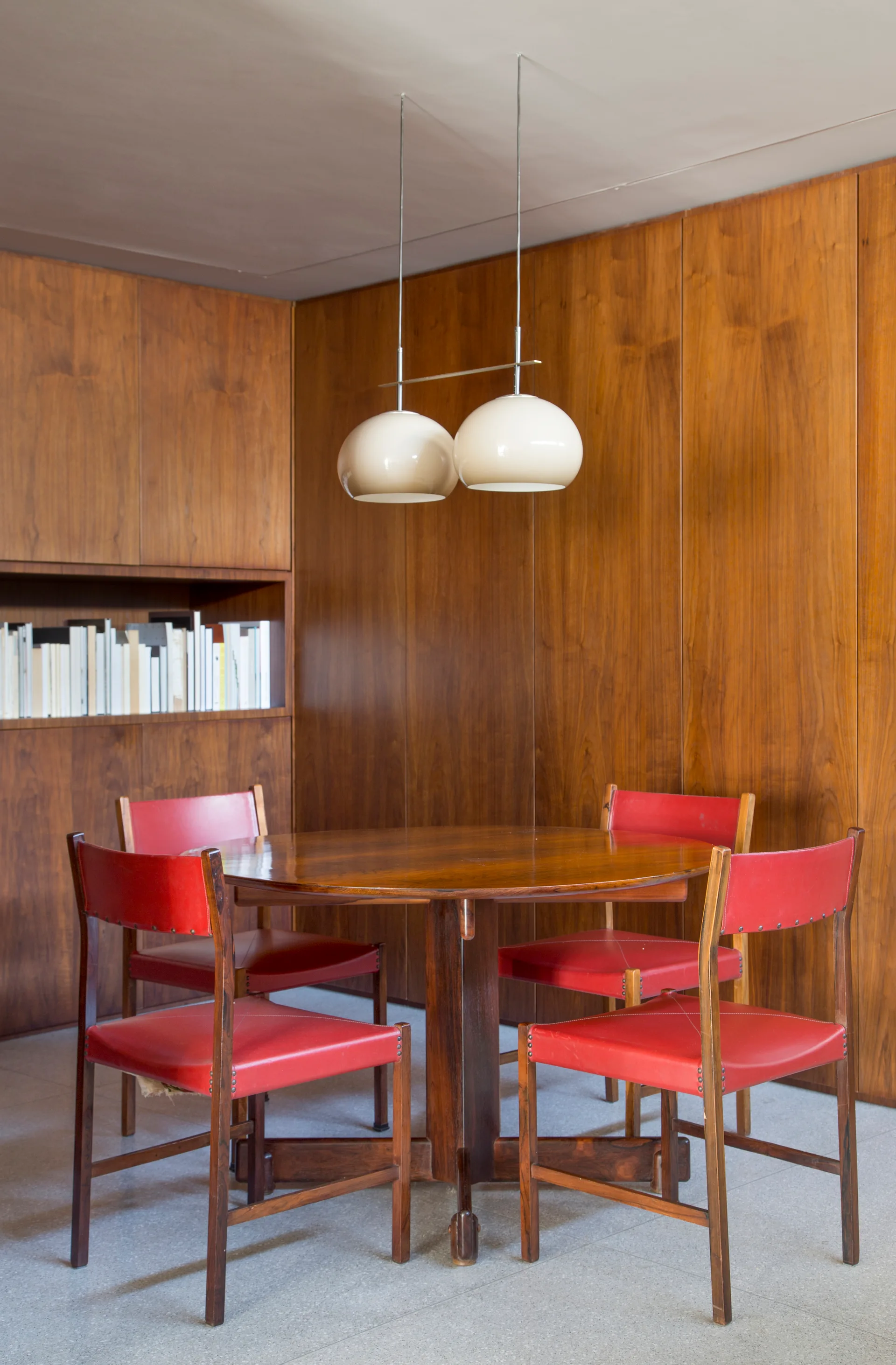 A round wooden dining table and four red leather chairs sit in a room with warm wood-paneled walls and a double dome pendant light.