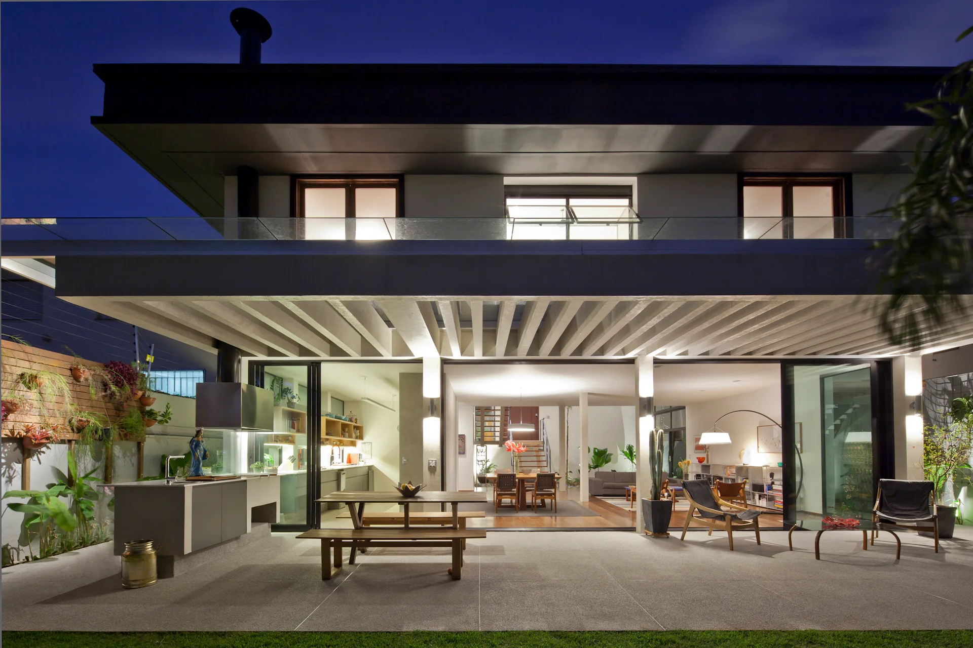 A modern covered patio features a built-in outdoor kitchen, a wooden dining table, and large sliding glass doors revealing an open-plan interior.