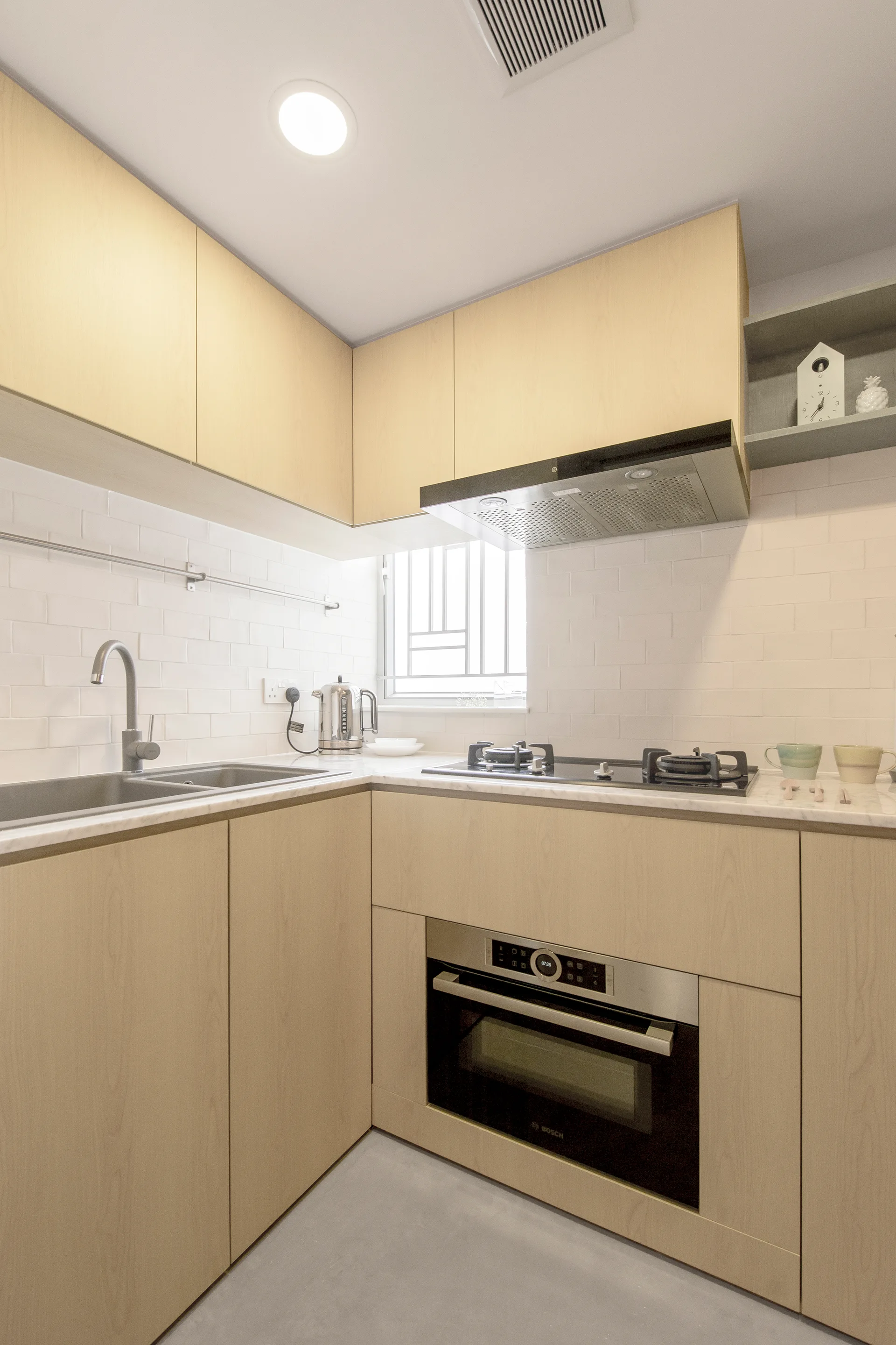 A compact L-shaped kitchen featuring light wood handleless cabinetry, a white subway tile backsplash, and an integrated stainless steel oven.