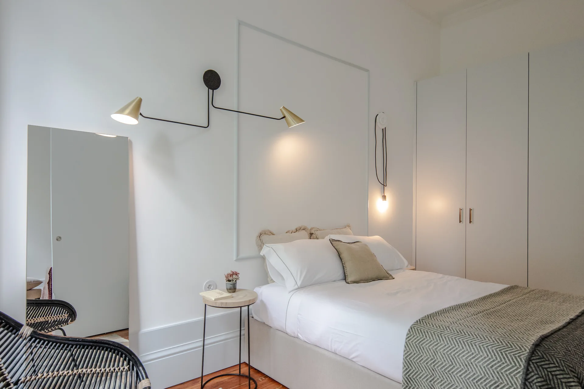 A minimalist white bedroom features an upholstered bed, a wall-mounted dual-arm brass sconce, wall molding, and white built-in wardrobes.