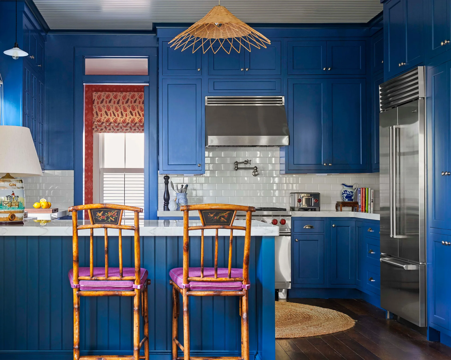 A kitchen features vibrant blue shaker cabinets, a white subway tile backsplash, a marble-topped island, and two rattan bar stools.