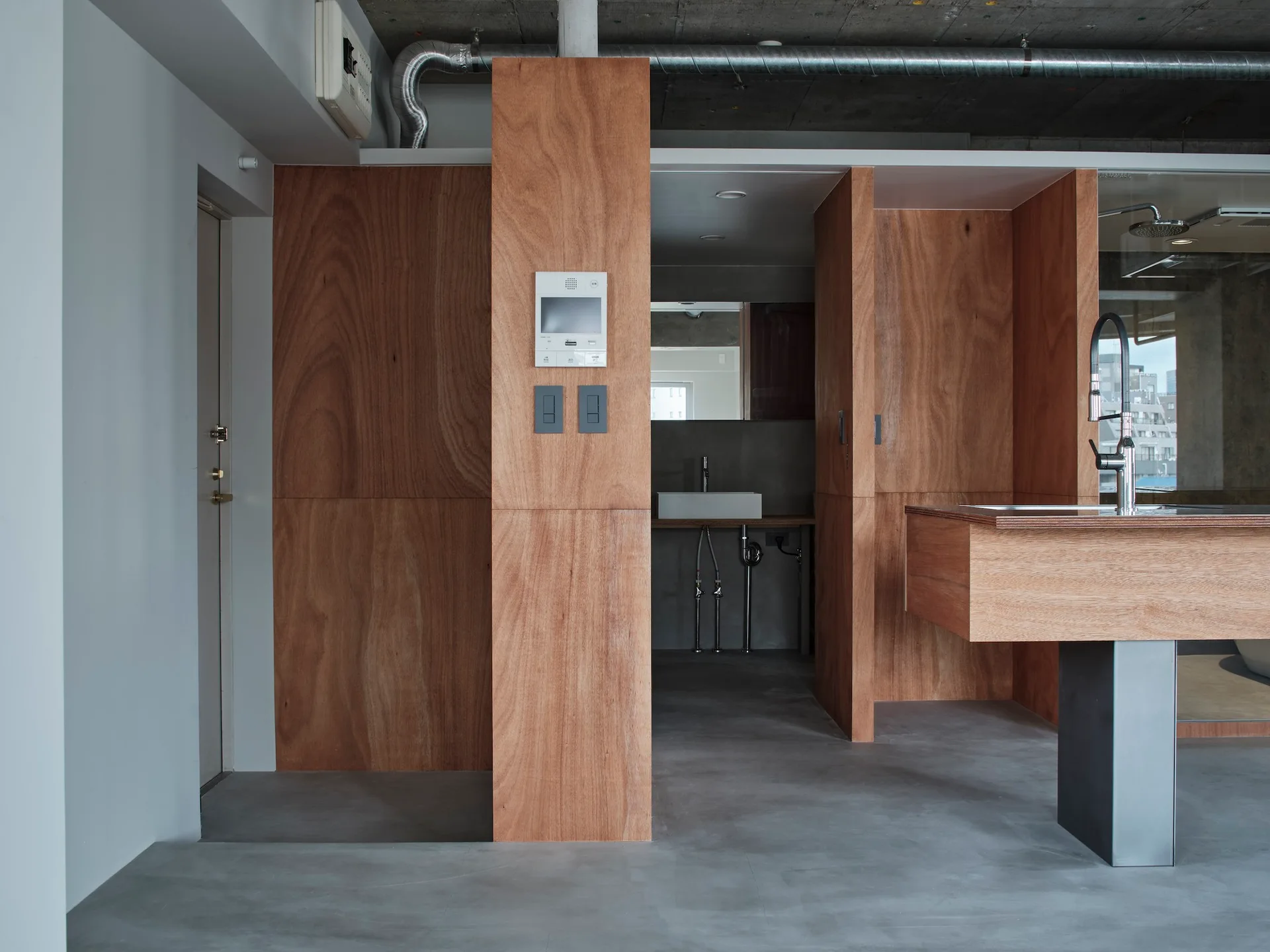 A minimalist industrial kitchen features warm plywood wall panels, a matching wood kitchen island with a steel faucet, and a grey concrete floor.