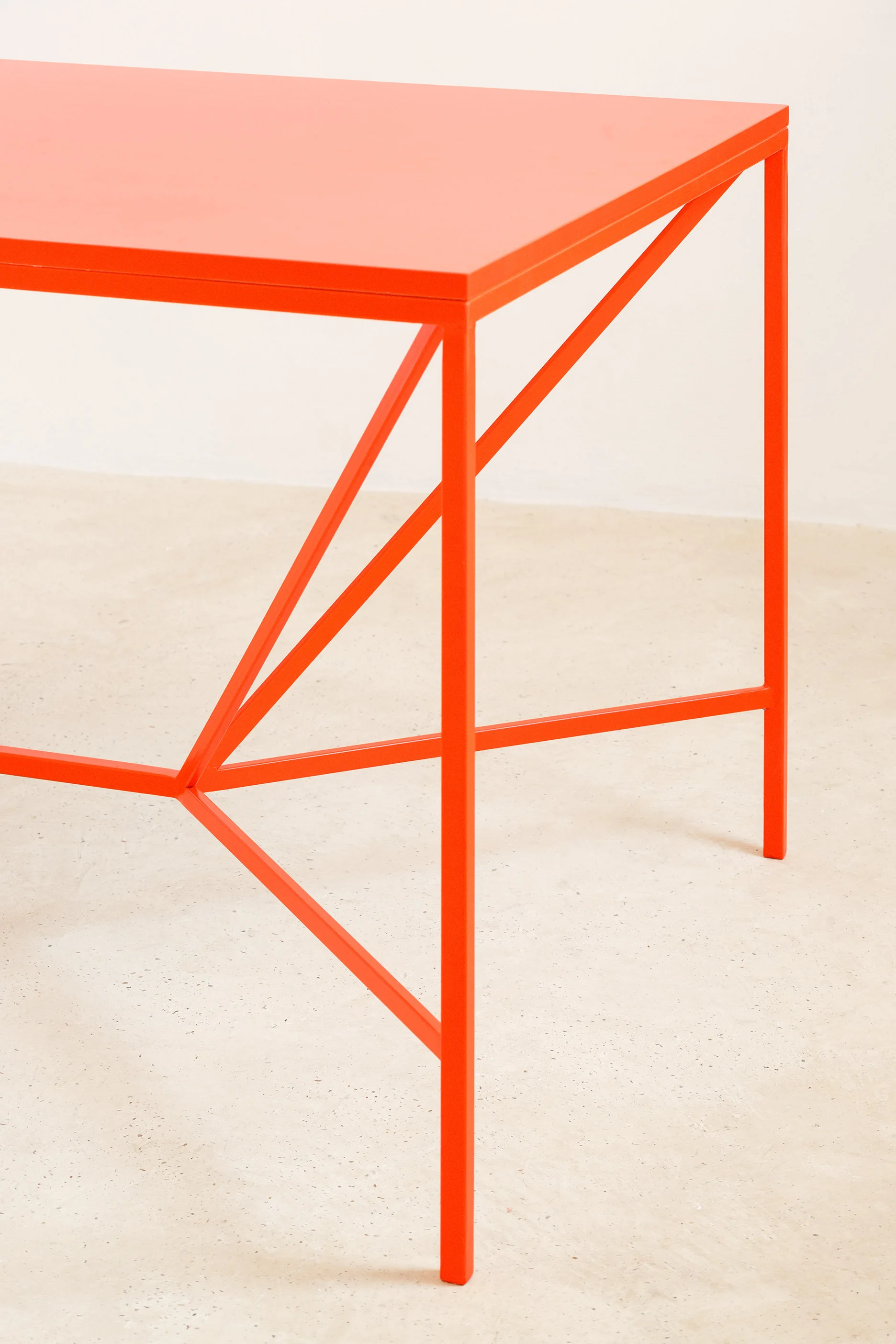 A bright orange metal table with a geometric leg base stands on a neutral speckled concrete floor against an off-white wall.