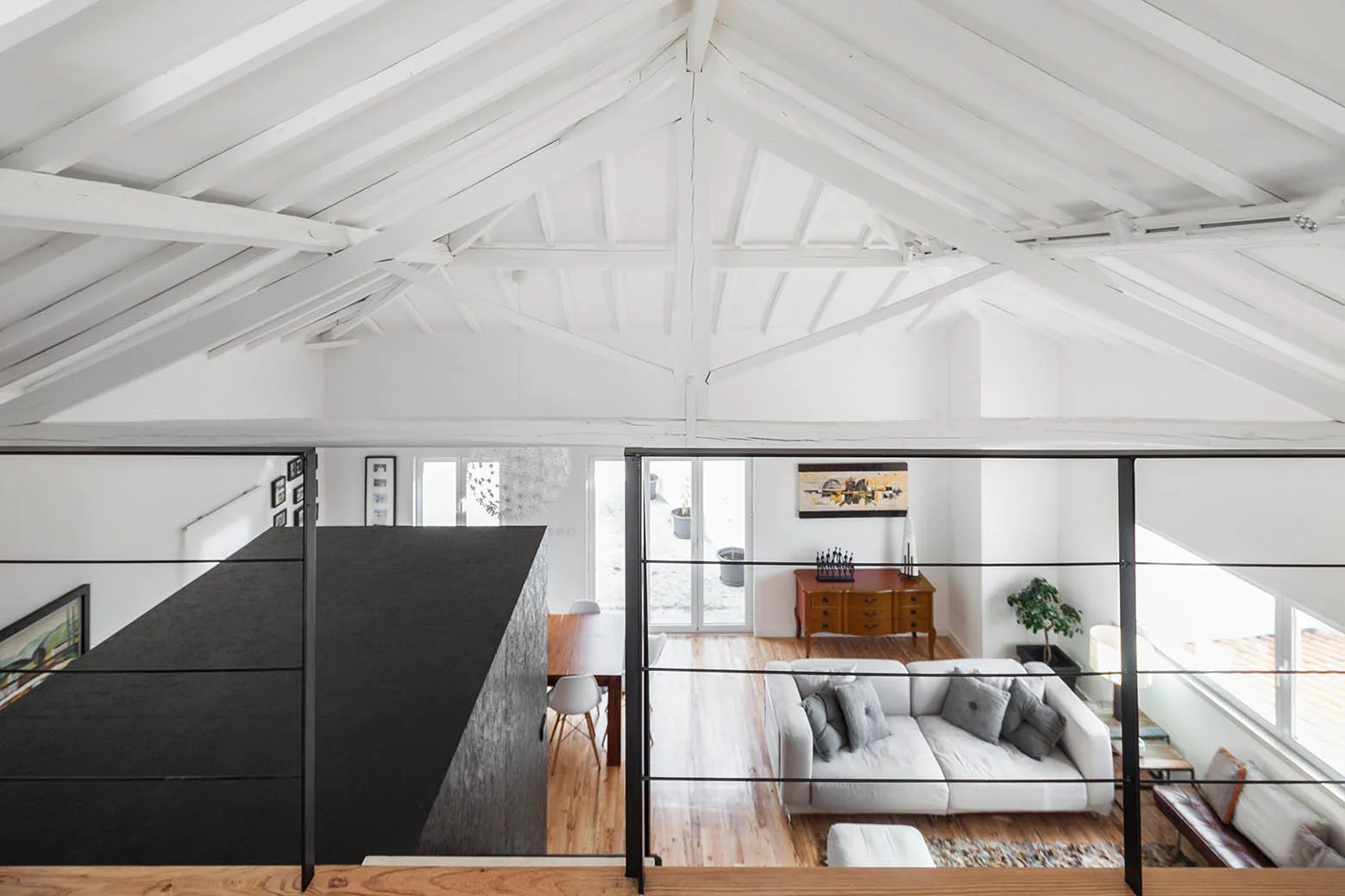 A view from a mezzanine with metal railings looking down at a living room with a white sofa, wood dining table, and white vaulted ceiling.