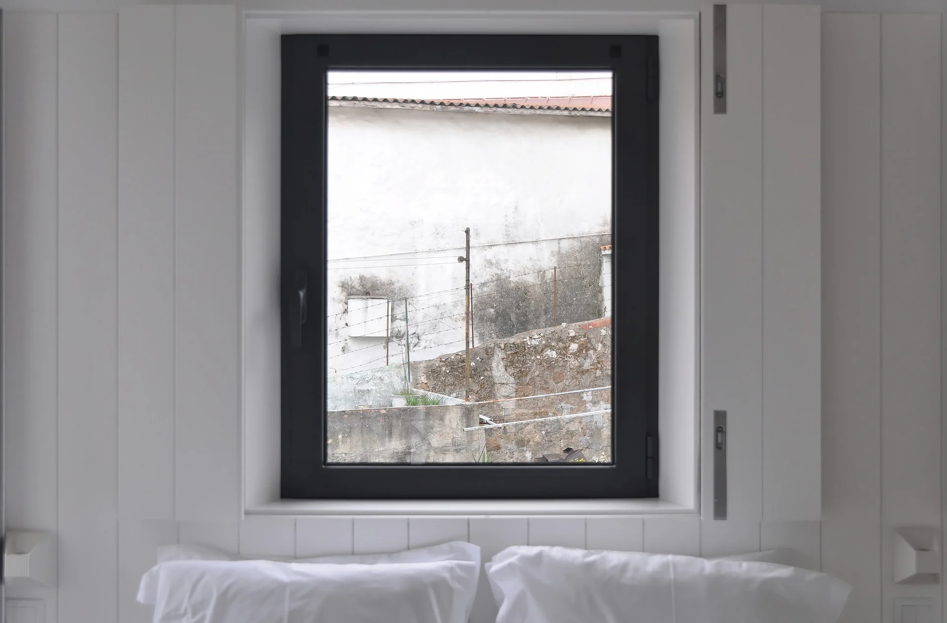A minimalist bedroom featuring white vertical wall paneling, a black-framed window, and two white pillows resting below.