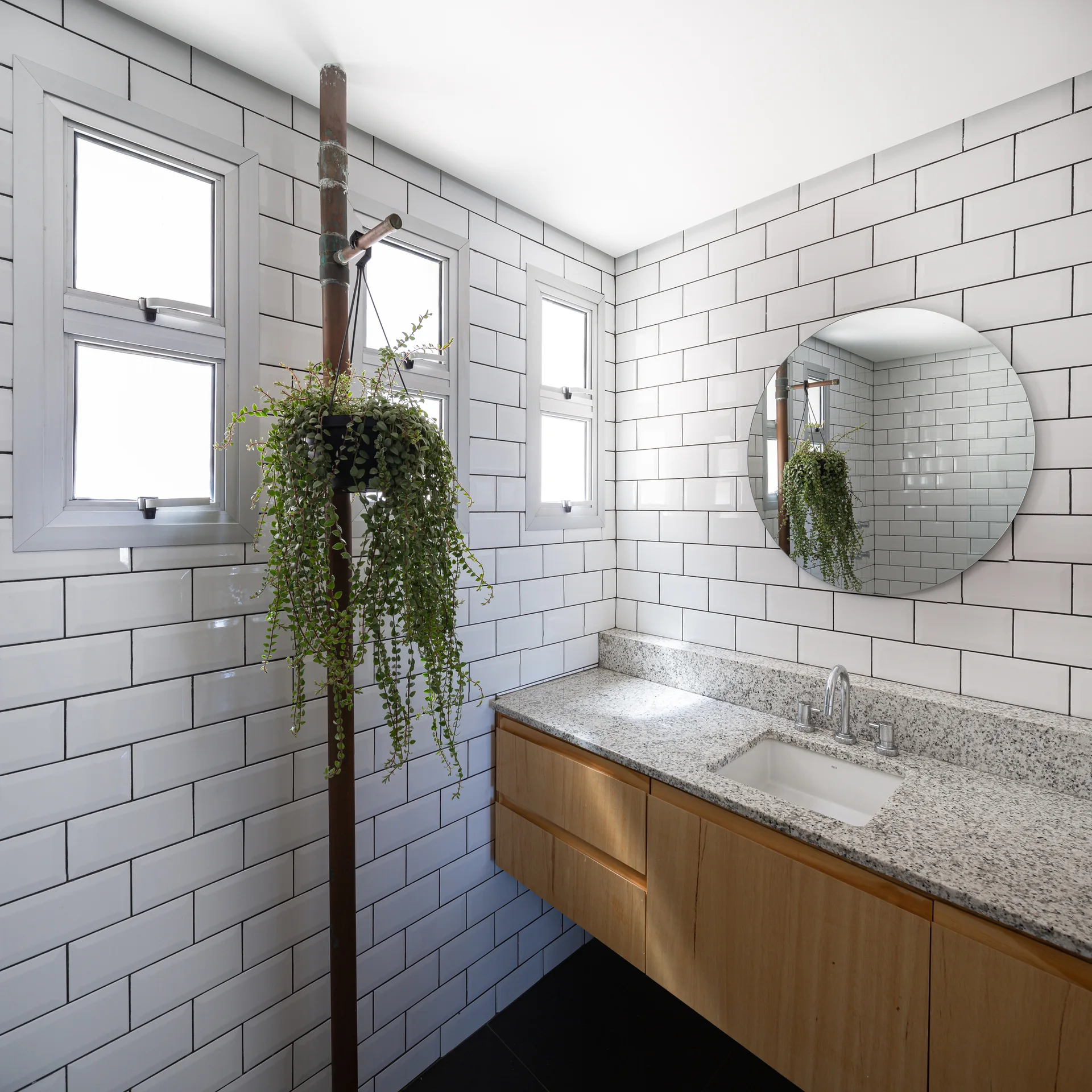 A modern bathroom features white subway tiled walls, a floating wood vanity with a granite countertop, a round mirror, and a hanging plant on a copper pipe.