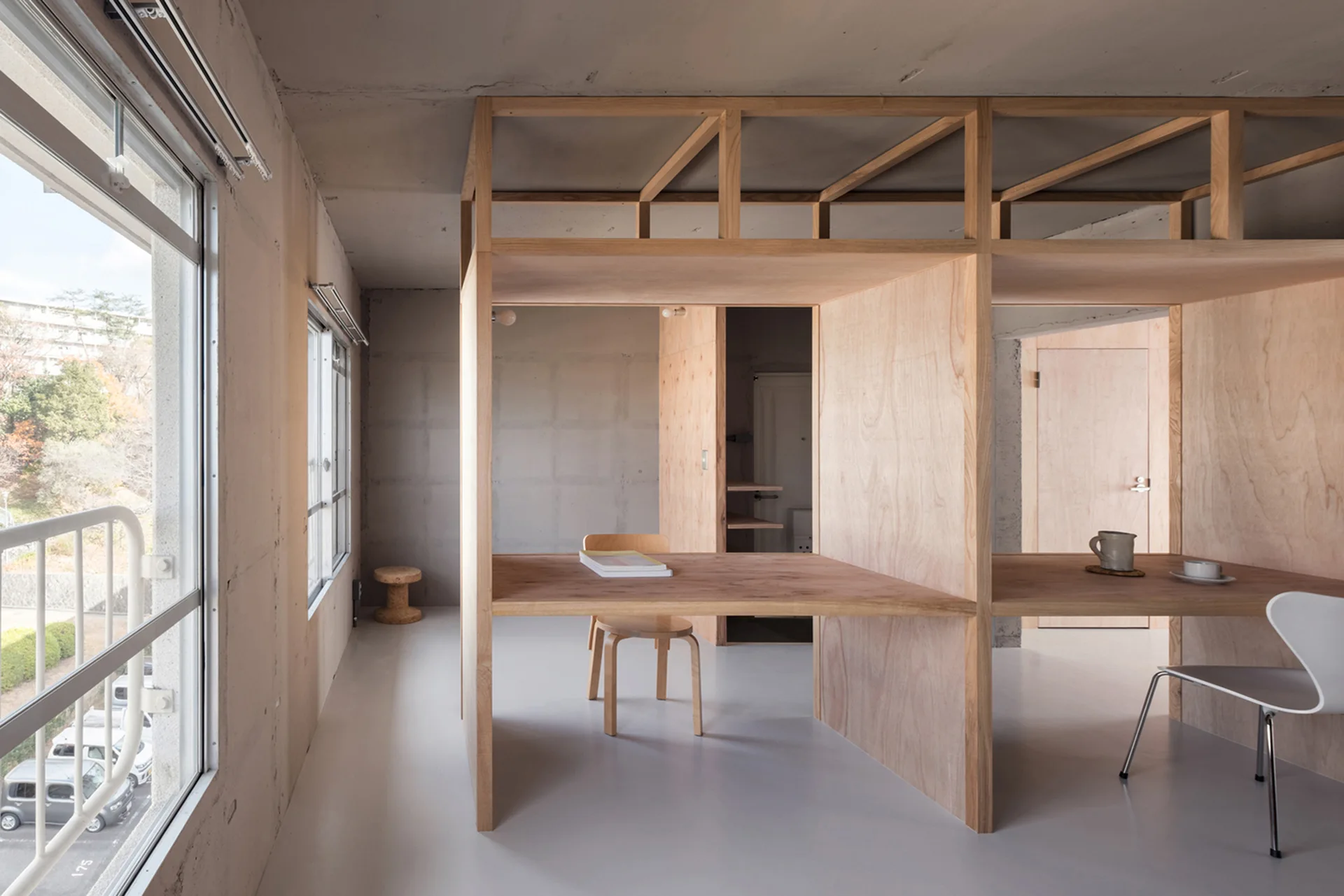 A minimalist home office featuring built-in wooden partitioned desks and chairs set against exposed concrete walls with large windows.