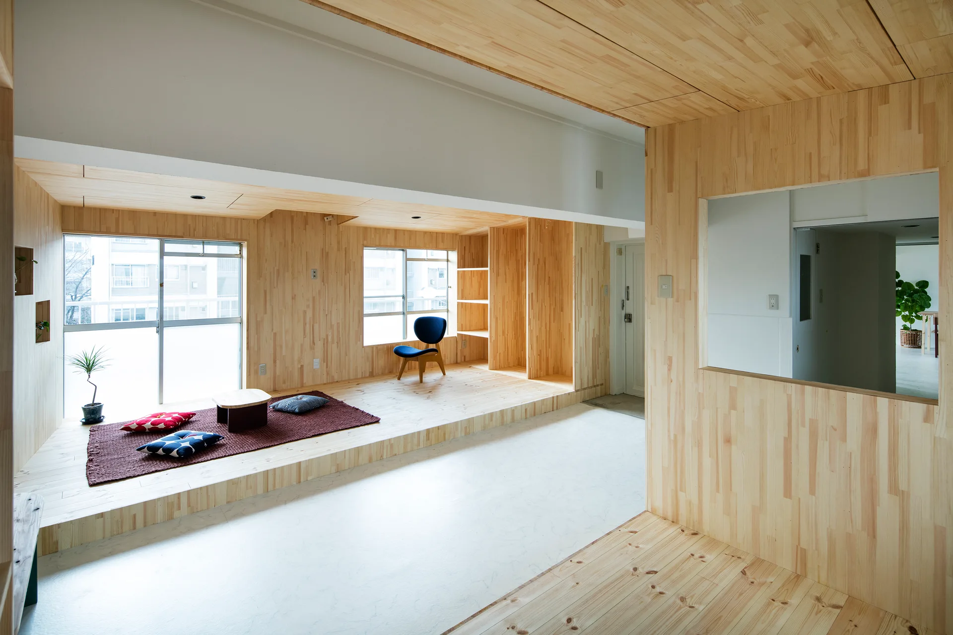 A residential room features a raised light wood platform with floor cushions, a low table, built-in shelving, and a blue chair.