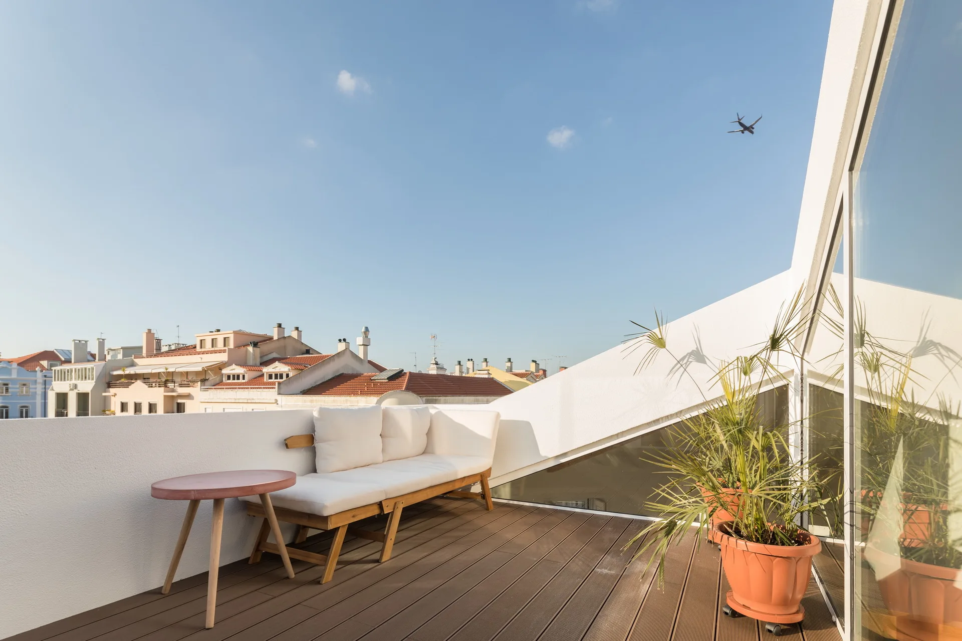 A sunny rooftop terrace features a wooden-framed sofa with white cushions, a pink side table, and terracotta planters on a dark wood deck.