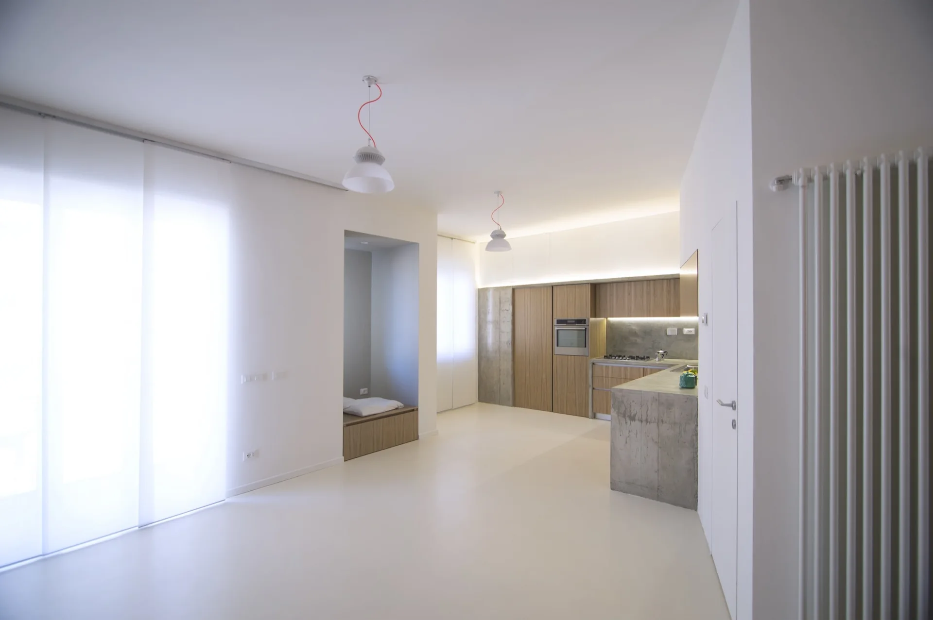 An open minimalist kitchen features a raw concrete island, wood-toned cabinetry, white floors, and a recessed wall niche with a bench.