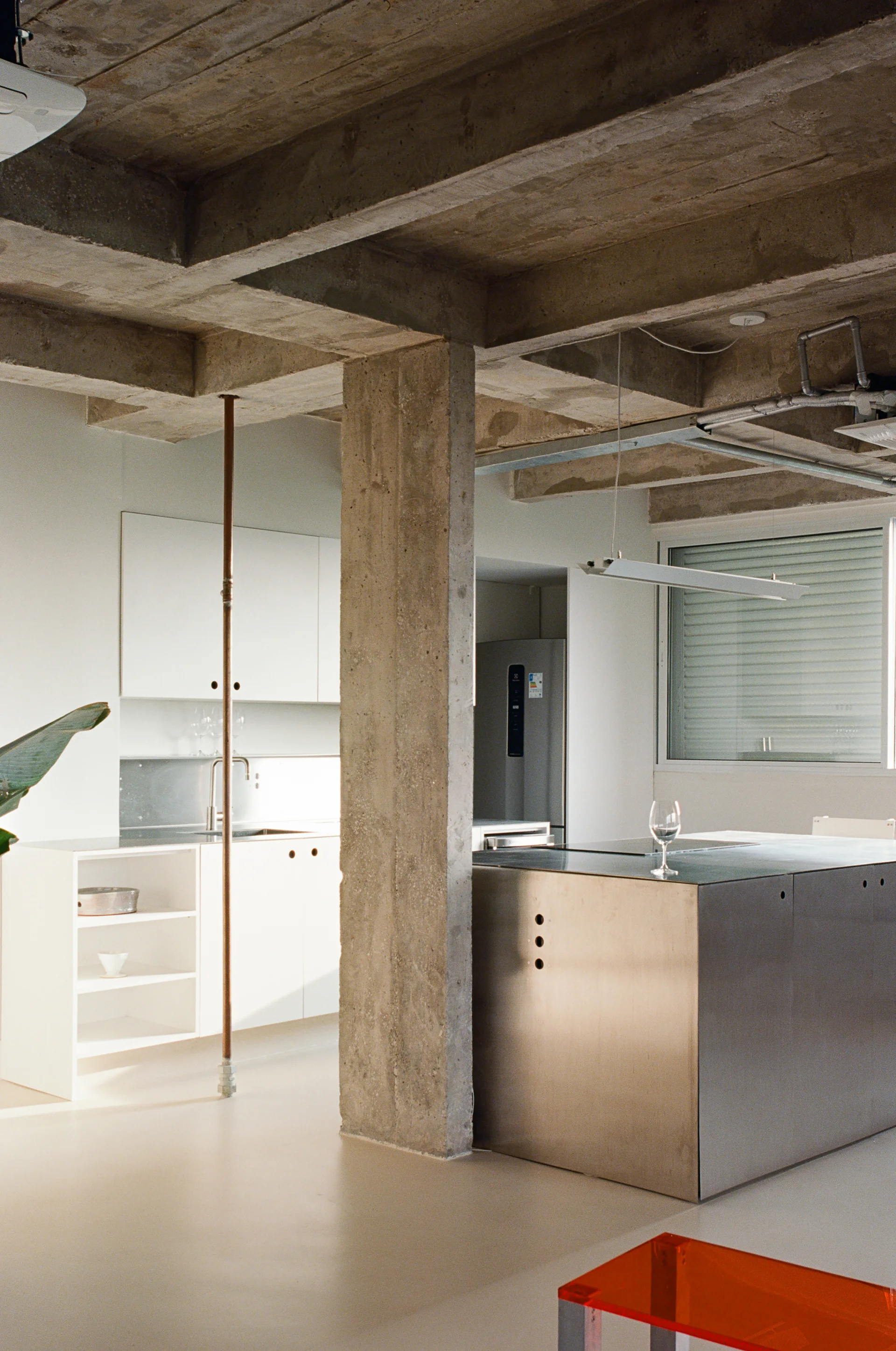 A minimalist kitchen features a large stainless steel island and white cabinetry set beneath an exposed concrete ceiling with a prominent structural column.
